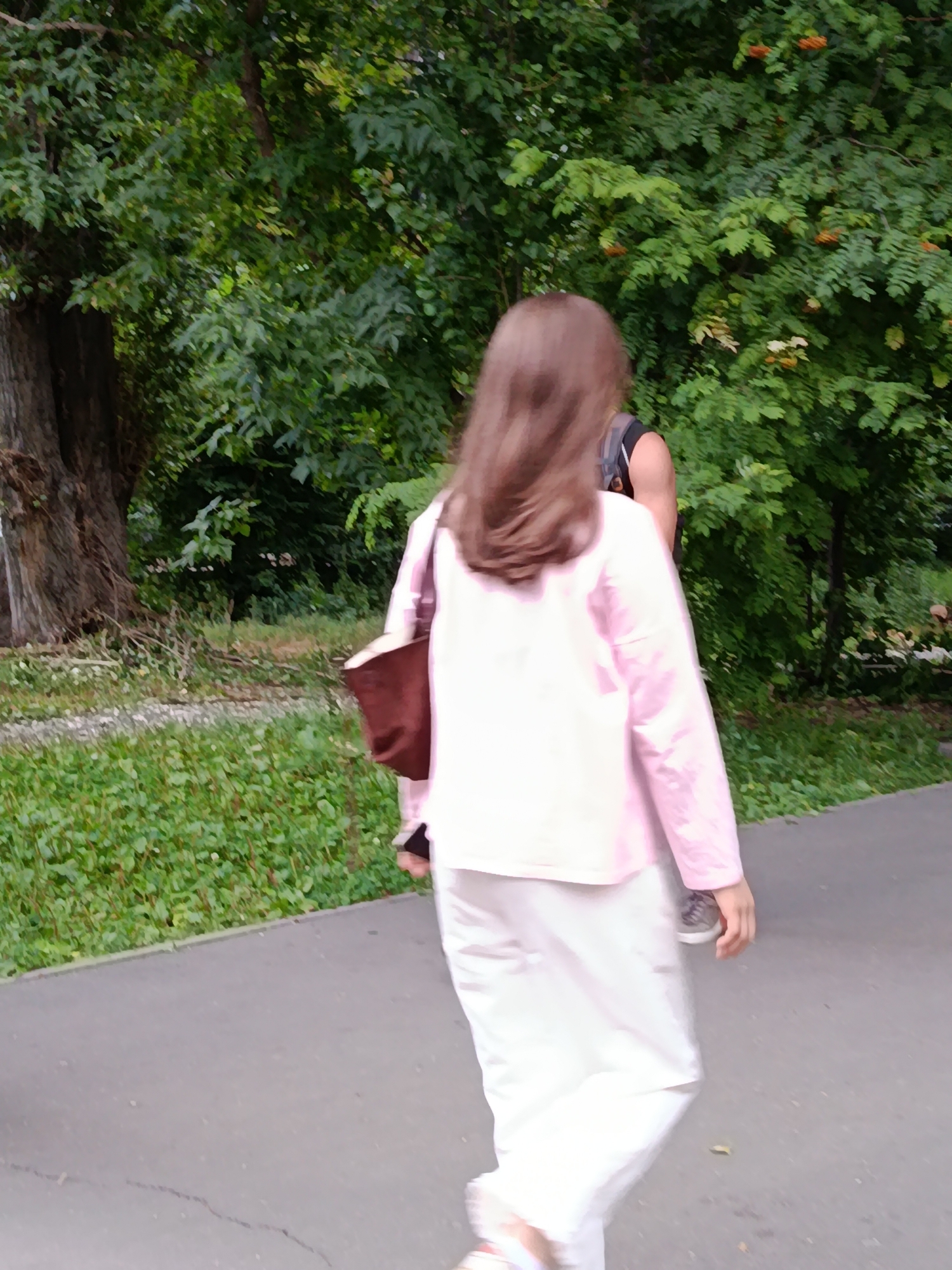 A person is walking away from the camera on a paved path in a park. The person has long brown hair and is wearing a light pink shirt and white pants. They are carrying a dark brown bag over their shoulder. There are trees and green foliage on either side of the path. The scene is outdoors during the daytime. The location is the city of Saratov, Russia.