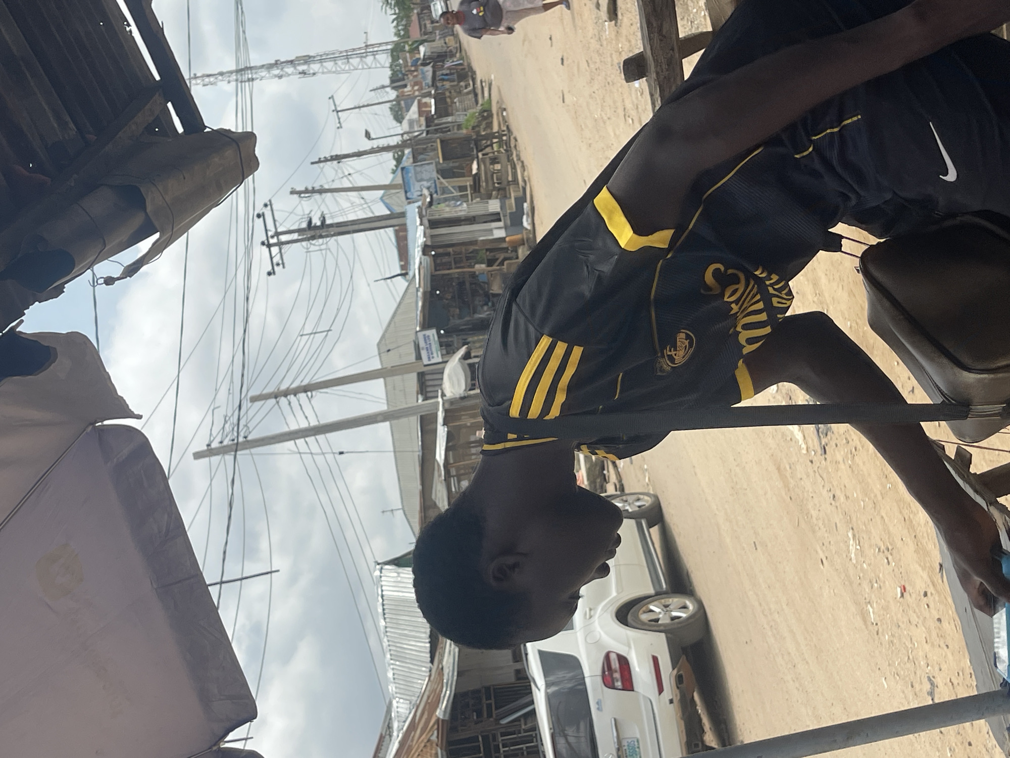 A street scene in Abuja, Nigeria. A dark-skinned young man in a black and yellow soccer jersey and black Nike shorts stands next to a metal structure. In the background are buildings and power lines, and a white SUV. Another person can be seen further down the street. The scene is in daylight under a cloudy sky.