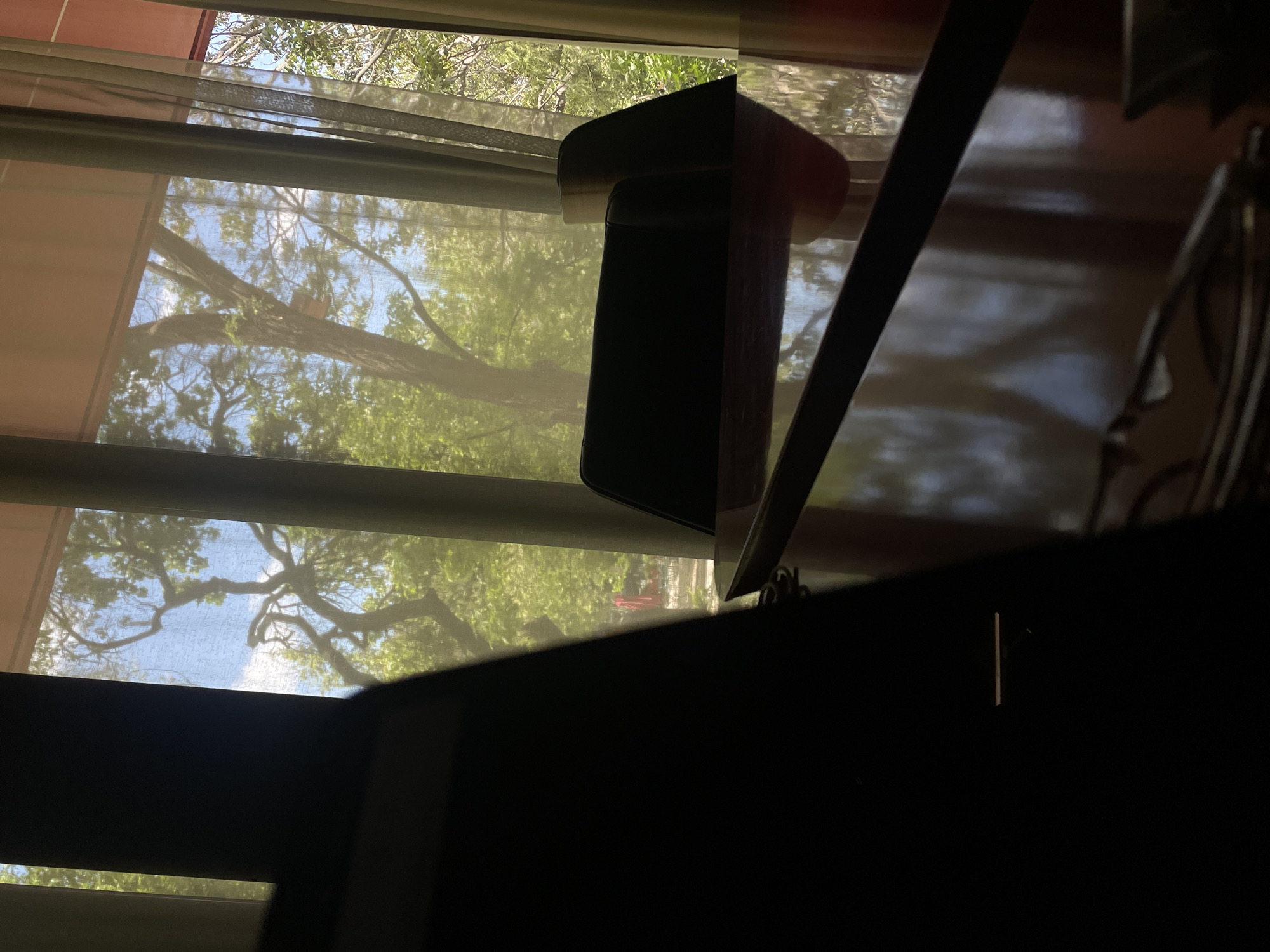 The image shows an interior view through a window. Outside the window, trees are visible with green foliage and a view of the sky. The window has a sheer curtain, which is partially obscuring the view.
In the foreground, there's a dark object, possibly furniture, which is silhouetted against the brighter outdoor view. A wooden object with a dark cylindrical part and a light-colored top is placed on top of the dark furniture. The lighting suggests the photo was taken during the day.
The location is unknown and cannot be determined from the information in the image. Therefore, it is not possible to confirm if it is in Yerevan, Armenia.