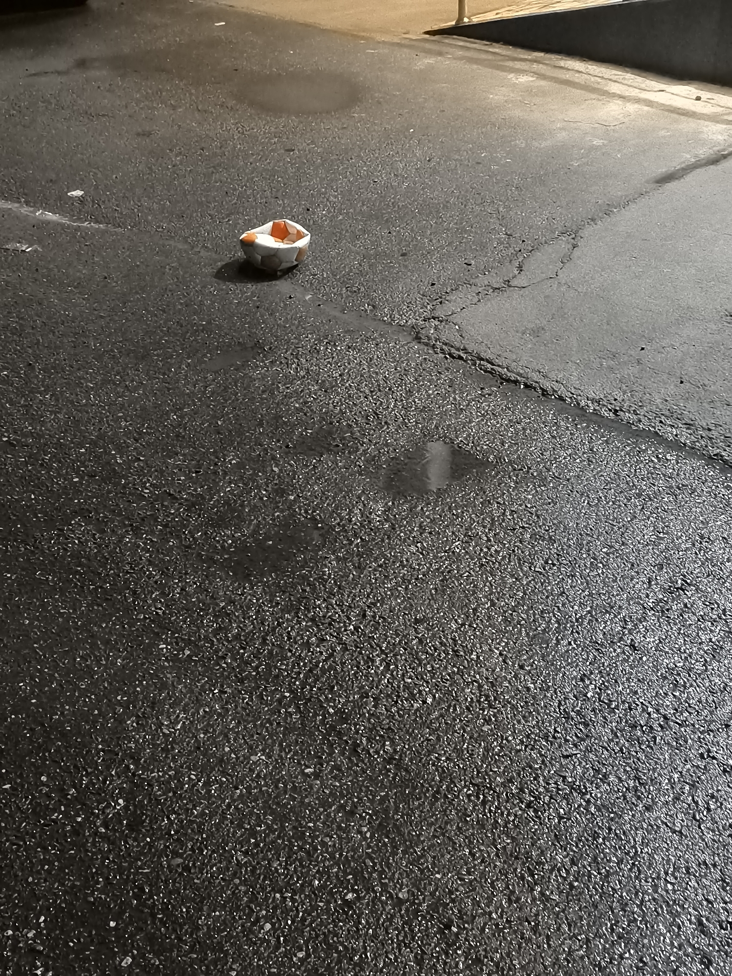 The image shows a discarded, crumpled soccer ball on a wet asphalt surface. The ball is mostly white with orange and black pentagonal panels. It appears to be deflated and misshapen, as if it has been run over or damaged. The asphalt is dark and wet, with several small puddles reflecting the dim light. There is a crack running diagonally across the surface, dividing it into two distinct areas. In the background, there is a blurred out area that appears to be a wall or building, with a hint of a tan or beige color. The lighting suggests it is either dusk, dawn, or nighttime. There is no visible text in the image. The scene is one of urban decay and neglect, with the damaged ball serving as a symbol of abandonment.