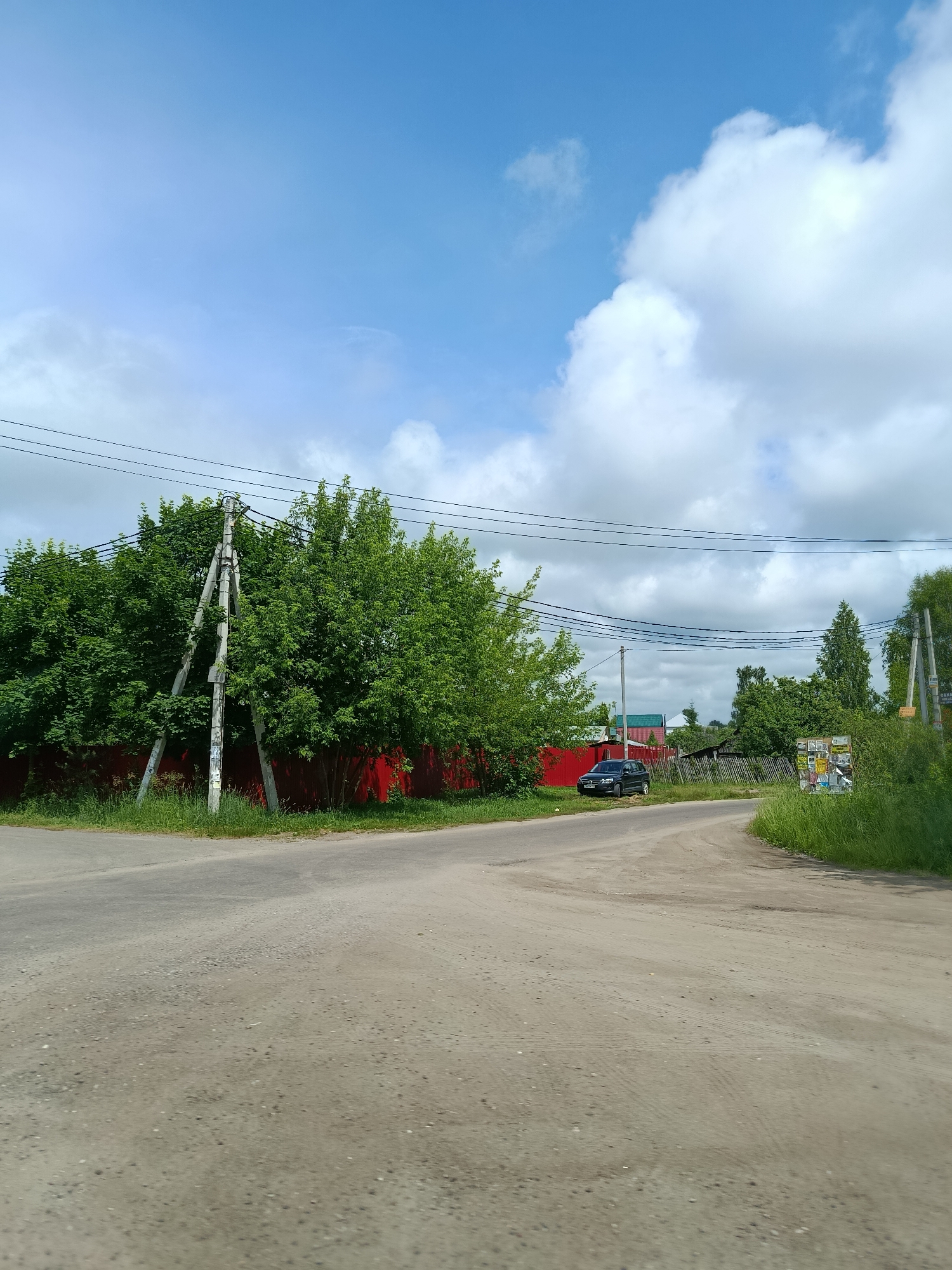 The image shows a view of a dirt road intersection in a rural area, possibly the city of Тополиный, Russia. A dark blue SUV is parked on the left side of the road. A bright red fence runs along the left side of the road, behind which are dense green trees. On the right side of the road, there is tall green grass and a wooden fence. Several utility poles and power lines are visible throughout the scene. The sky is blue with scattered white clouds. The overall scene depicts a quiet, possibly residential, area.