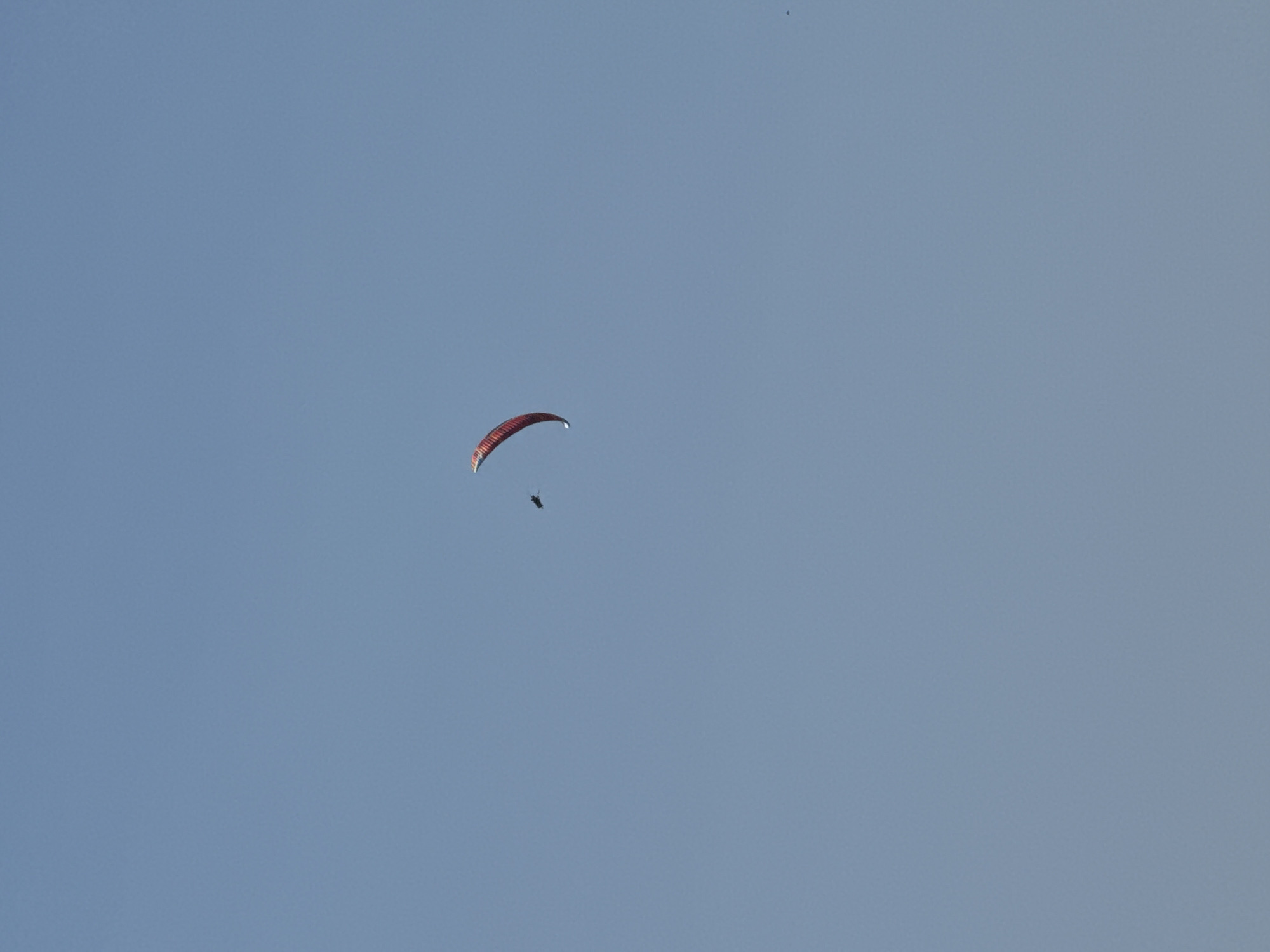 In the expansive, clear blue sky above Boreti, Montenegro, a lone kitesurfer is captured mid-air. The kitesurfer, a small dark silhouette against the bright sky, is attached to a vibrant red kite that curves gracefully in the wind, forming a crescent shape. The kite, a striking splash of color, dominates the upper left quadrant of the image. The overall scene is one of open space and airborne activity, with the vastness of the sky serving as the backdrop.