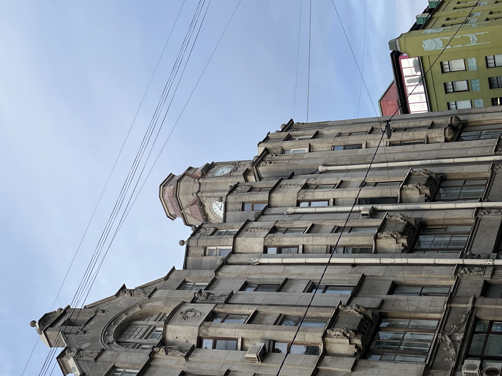 An ornate grey stone building, characteristic of early 20th-century architecture, dominates the frame, captured from a low, tilted angle looking upwards. The building features a stepped or terraced facade with numerous windows, many of which appear to be set back. Rich decorative elements, including sculpted figures, corbels, and intricate window frames, adorn the structure. Two clocks are visible on the upper section of the building; one clearly shows approximately 10:10, and the other, partially obscured, appears to be around 10:05. Several air conditioning units are mounted on ledges. White drainage pipes run horizontally across sections of the facade.
In the upper right background, a contrasting light green building with white decorative relief patterns is visible, suggesting it's situated behind or next to the main building. A reddish structure is briefly seen between the two buildings. The sky is a clear, light blue with minimal cloud cover, indicating good weather. Multiple thin black utility or tram wires stretch diagonally across the sky, intersecting the buildings. There are no people visible, and no specific event is taking place; the scene is static. The time of day is morning, as indicated by the clocks. Based on the provided context, the location is Saint Petersburg, Russia. No legible text is visible on any of the structures.