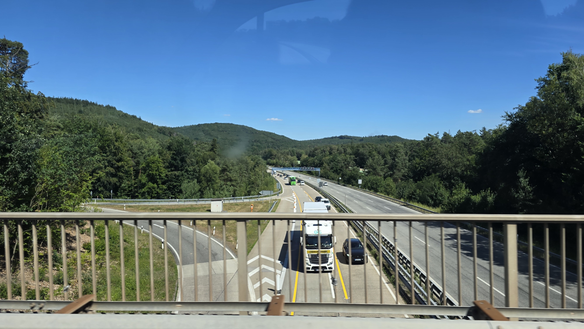 This is a photograph of a highway in Kaiserslautern, Germany, on a clear day with a blue sky. The perspective is from an elevated viewpoint, likely a bridge over the highway, with a metal railing in the foreground. The highway itself is multi-lane and appears busy with traffic. A white truck with a yellow stripe on its side is prominent in the center of the frame, traveling in the right lane. A black car is to the right of the truck. Other vehicles, including cars and a green bus, are visible further down the highway. The landscape surrounding the highway is characterized by lush green, tree-covered hills. The overall scene depicts a typical German autobahn cutting through a forested area.