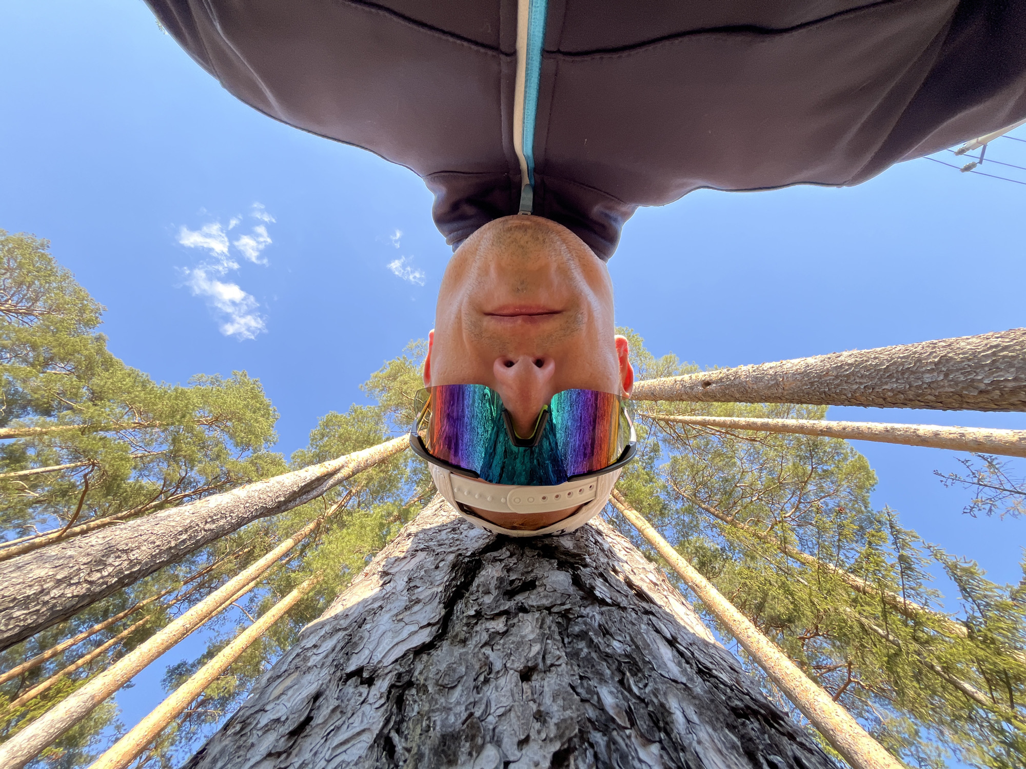 A man is seen upside down in the photograph, positioned against a tree trunk. He's wearing a dark jacket with a light blue stripe, a white baseball cap, and reflective sunglasses. The background consists of tall pine trees reaching into a blue sky with a few white clouds. The perspective suggests the photo was taken from a low angle, looking upwards. There is no indication of the city of Chaikovsky, Russia.