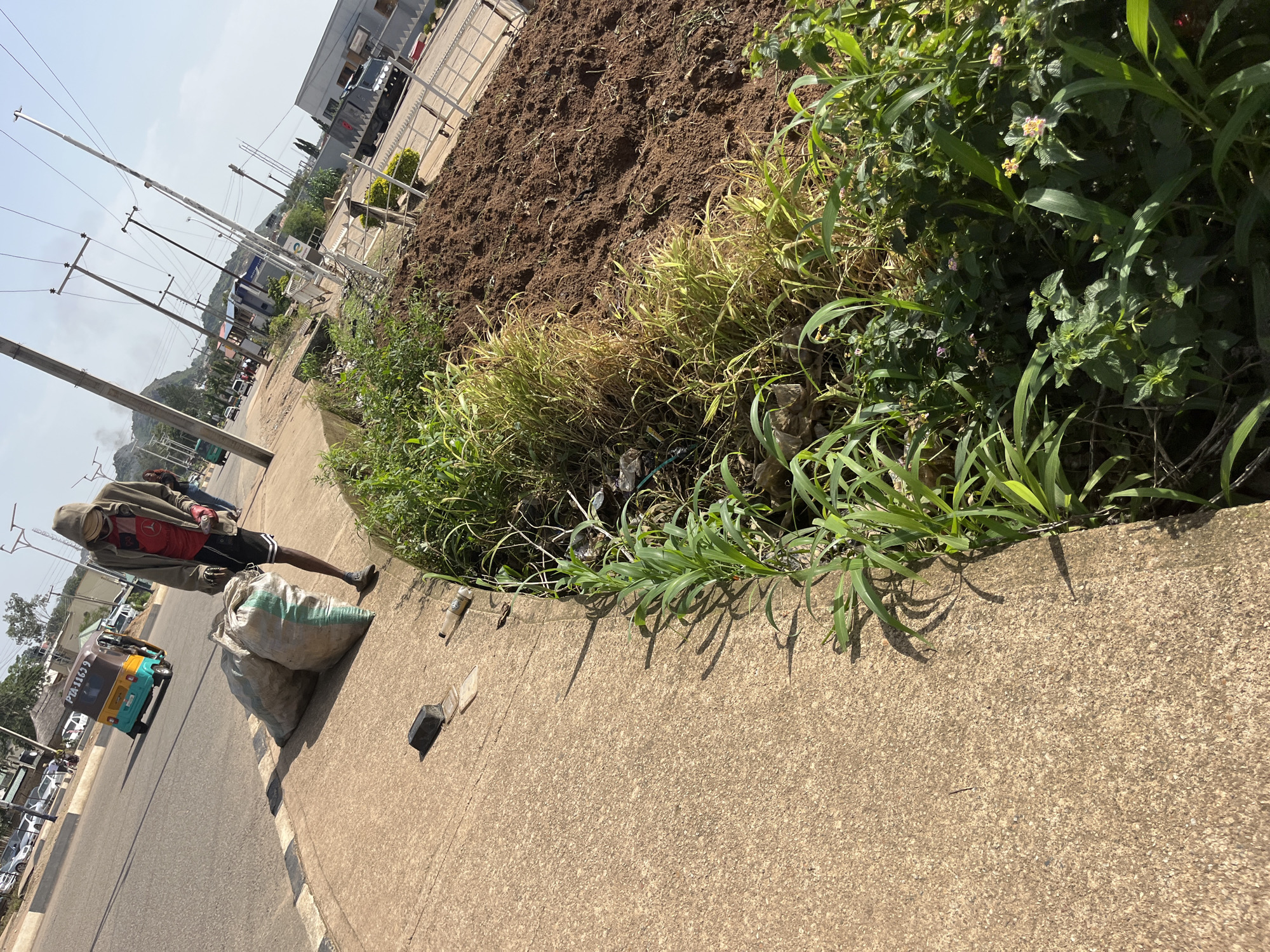 In Bukuru, Nigeria, a person wearing a hooded jacket, a red shirt, black shorts, and sneakers stands on a concrete sidewalk next to a dirt patch. They are carrying two large, light-colored sacks. A yellow and blue tricycle is visible on the road in the background, along with other vehicles and utility poles. The scene appears to be along a street with vegetation and construction or landscaping in progress.