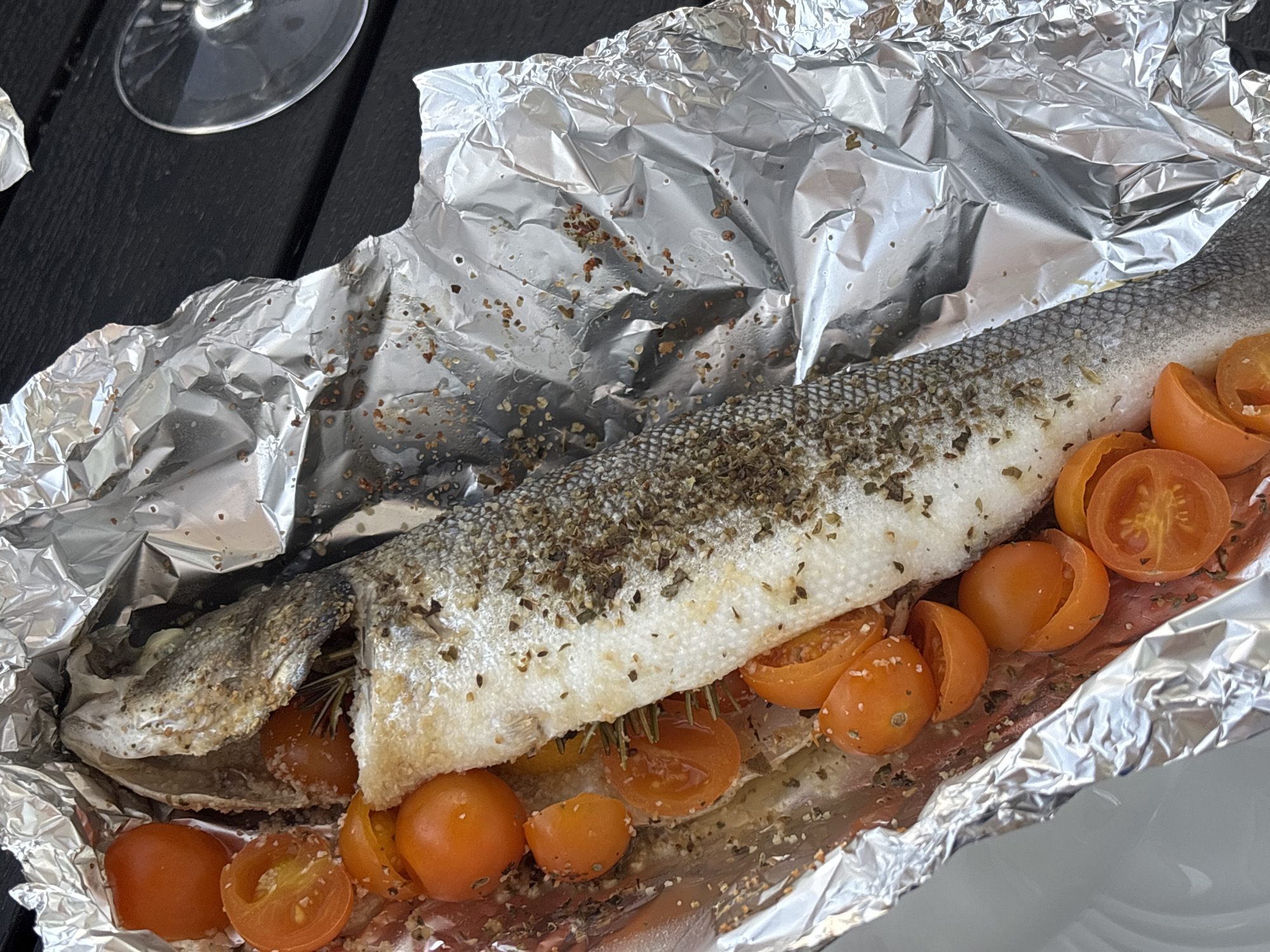 An overhead shot shows a whole fish baked in foil with cherry tomatoes. The fish is seasoned with herbs and spices. The foil is shiny and crinkled, and the tomatoes are halved and scattered around the fish. The meal is presented on a dark, textured surface, possibly a wooden table. In the background, a clear wine glass is visible, suggesting an outdoor dining setting. The image is well-lit, with natural light highlighting the textures and colors of the food. The scene is likely a casual meal enjoyed outdoors. Without further context, the specific location in Tavira, Portugal cannot be determined from the visual information provided.