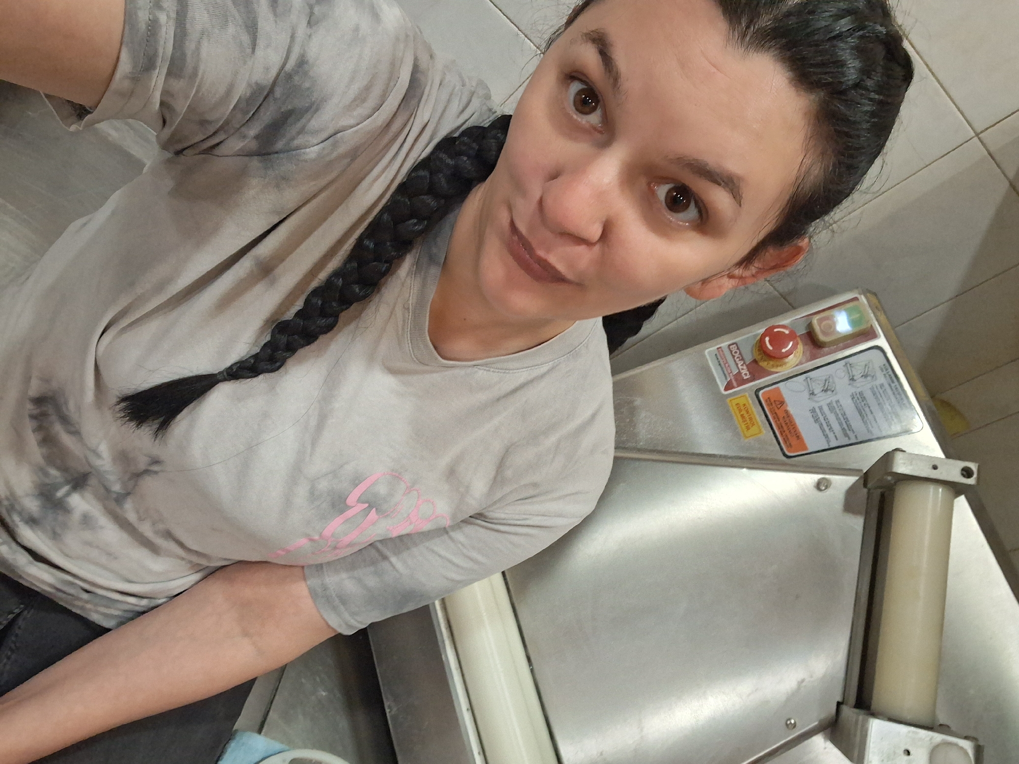 A person with dark hair styled in a braid and wearing a grey tie-dye t-shirt is standing next to a piece of industrial machinery. The machine has a metal surface and a control panel with a red emergency stop button and a green on/off switch. Warning labels and instruction diagrams are affixed to the machine. The location and action details are indeterminable. The city and country context is not discernible from the image.