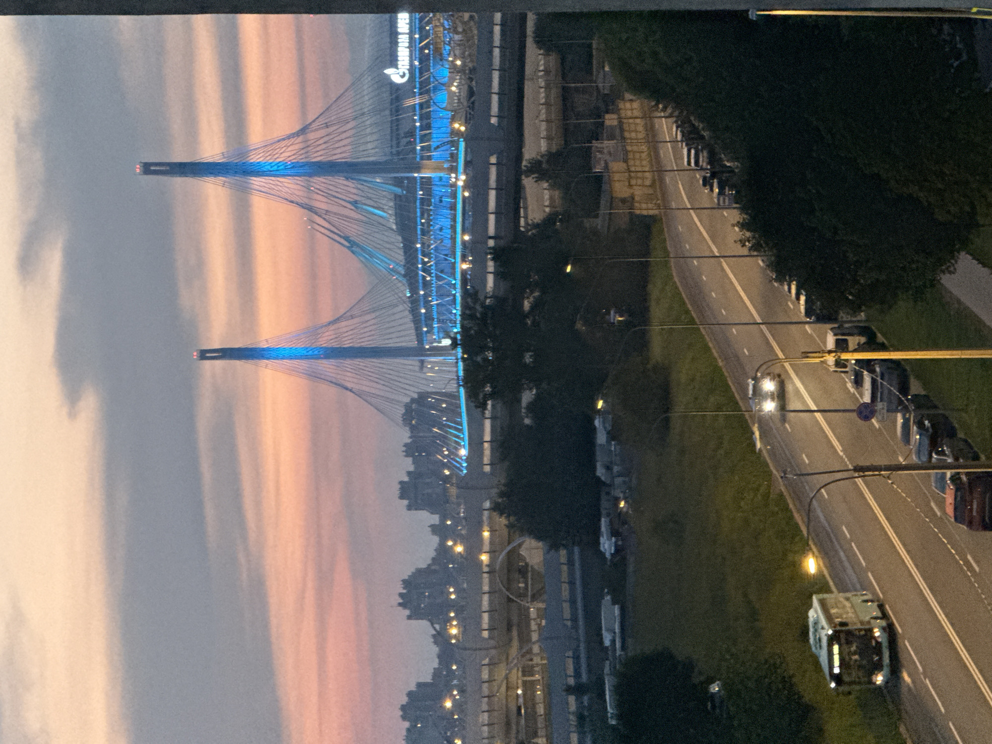 The image captures an evening view of the Gazprom Arena stadium in Saint Petersburg, Russia, with its distinctive cable-stayed bridge illuminated in blue. The stadium is located on Krestovsky Island, near the Gulf of Finland. In the foreground, a two-lane road curves through a green, grassy area. A light blue bus is traveling on the left lane of the road, its headlights and interior lights on. Several cars are parked along the right side of the road, and two cars are moving in the opposite direction in the right lane. The sky is a soft gradient of pink and gray, indicating dusk or dawn, with clouds obscuring the horizon. Distant buildings, also illuminated, can be seen behind the stadium and bridge.