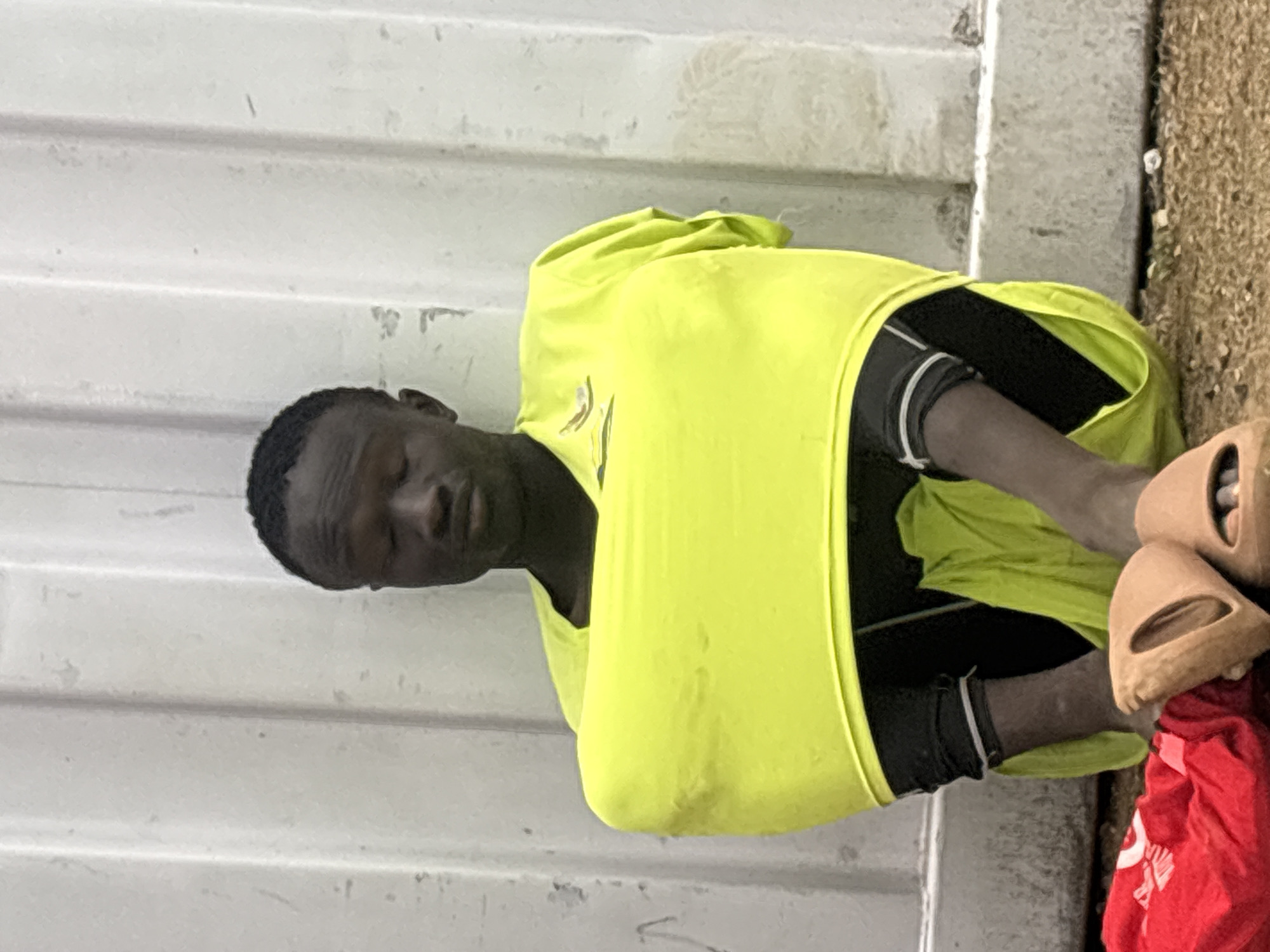A man wearing a bright yellow shirt, black shorts, and light-colored sandals is crouched against a corrugated metal wall in what appears to be an outdoor location in Bukuru, Nigeria. His eyes are closed and he is holding his legs. A red cloth or bag is visible on the ground next to him. The surrounding environment appears to be a dusty or sandy area.
