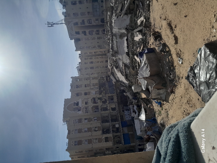 The image depicts a scene of urban destruction and temporary settlement in Khan Yunis, Palestinian Territory, under bright sunlight. In the background, a row of multi-story residential buildings, primarily light beige, exhibits extensive structural damage, characterized by numerous missing windows, damaged balconies, and large holes in their facades. One central building features a prominent arched architectural element. A cellular communication tower is mounted on the roof of a building on the far right.
Below these damaged structures, a large area is filled with demolition debris, including shattered concrete slabs, twisted rebar, and general rubble, indicating collapsed buildings. In the midground, on an expanse of sandy, disturbed earth, a cluster of approximately ten makeshift tents is erected. These temporary shelters are constructed from various materials such as tarpaulins and fabric sheets in shades of white, brown, and blue. Some tents display attached banners or signs. A few indistinct human figures are visible on the far left, near the tents and damaged structures, their specific actions not discernible. In the immediate foreground, a light blue fuzzy fabric is draped over a white ledge, partially obscuring a black plastic sheet on the ground below. The sky is clear and bright.