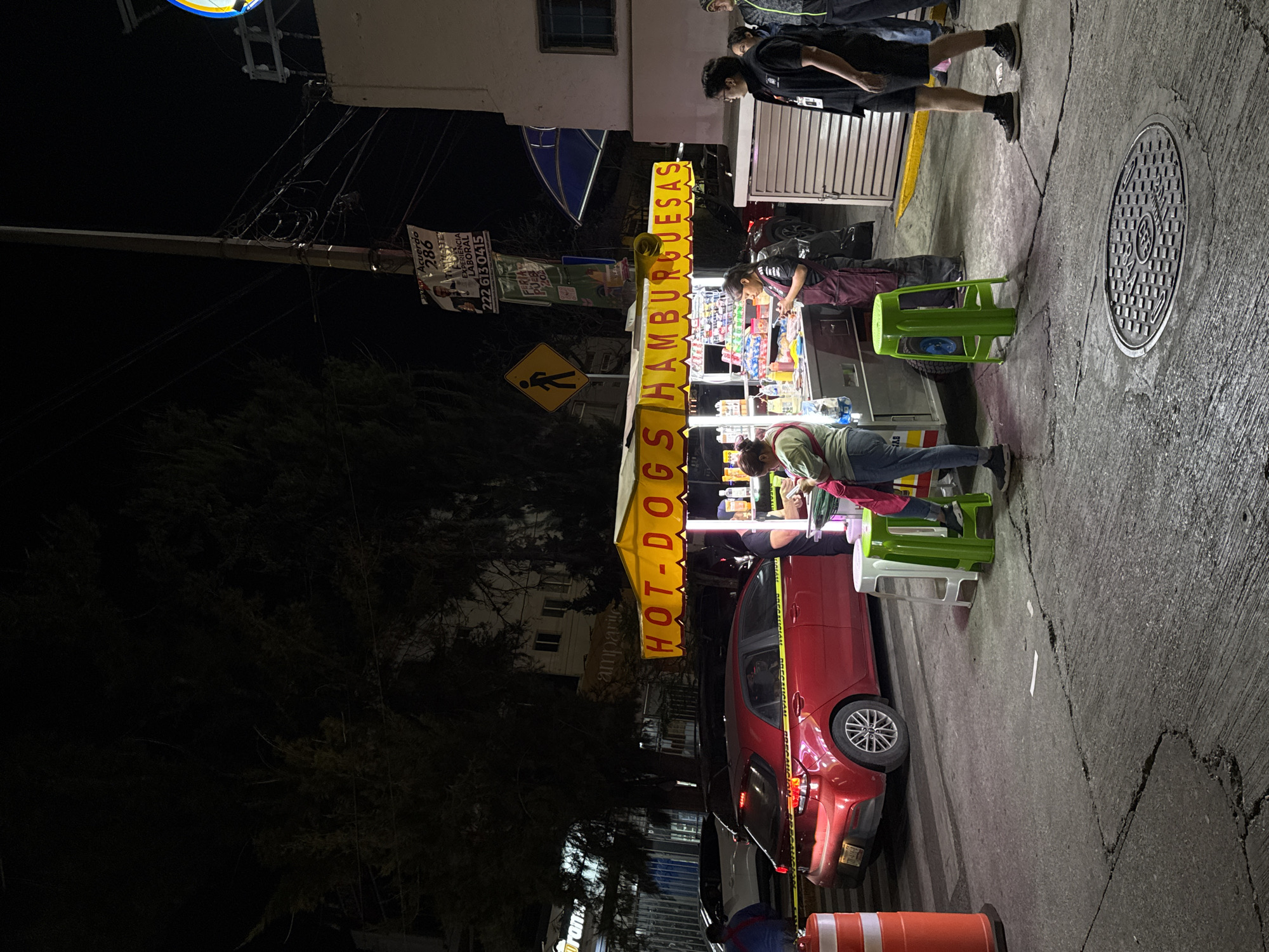 The image captures a street scene at night, likely in Puebla, Mexico, given the presence of Spanish signage and cultural elements. A brightly lit food cart, with "HOT-DOGS HAMBURGUESAS" in neon signage, is the focal point. Customers are interacting with the food cart. A red hatchback car is parked adjacent to the cart, with caution tape indicating a possible incident. Pedestrians are walking along the sidewalk. A manhole cover is visible on the sidewalk. A pedestrian crossing sign is visible. A political poster is fixed to a street lamp. The left side of the image is dominated by darkness and trees.