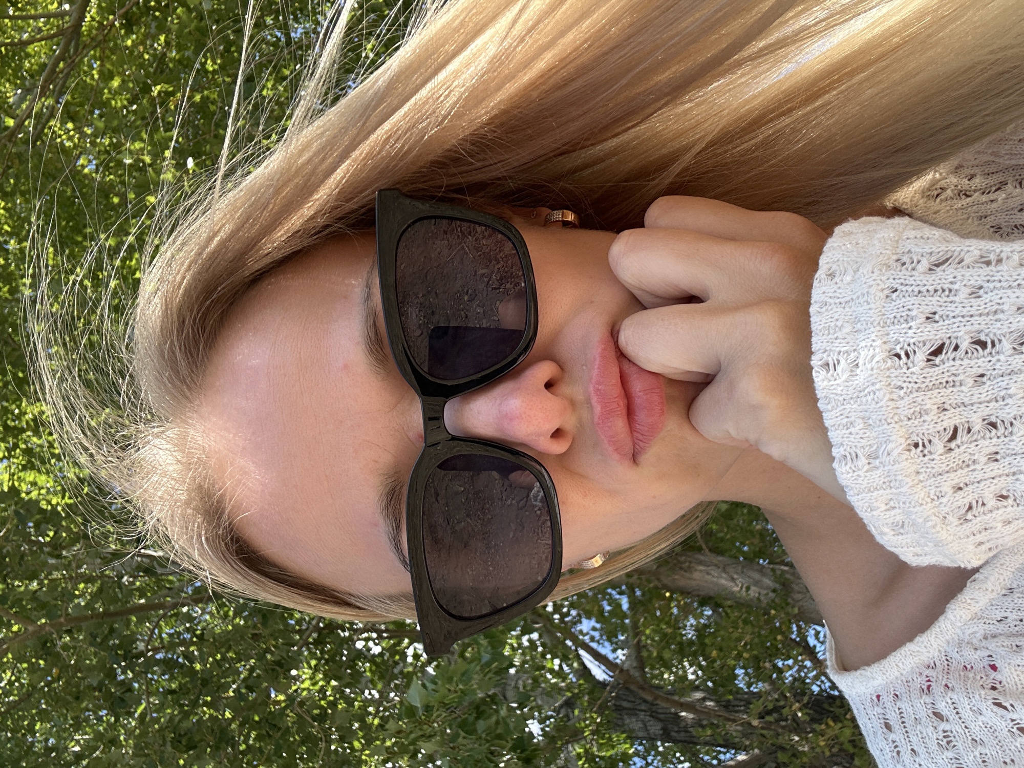 A close-up, head and shoulders portrait of a young woman with blonde hair and sunglasses is taken outdoors against a backdrop of green trees and dappled sunlight. The woman is resting her chin on her hand, and her mouth is pursed. She is wearing a white, loosely knit sweater. The scene is likely in Gornyi Erik, Russia, given the environmental context.