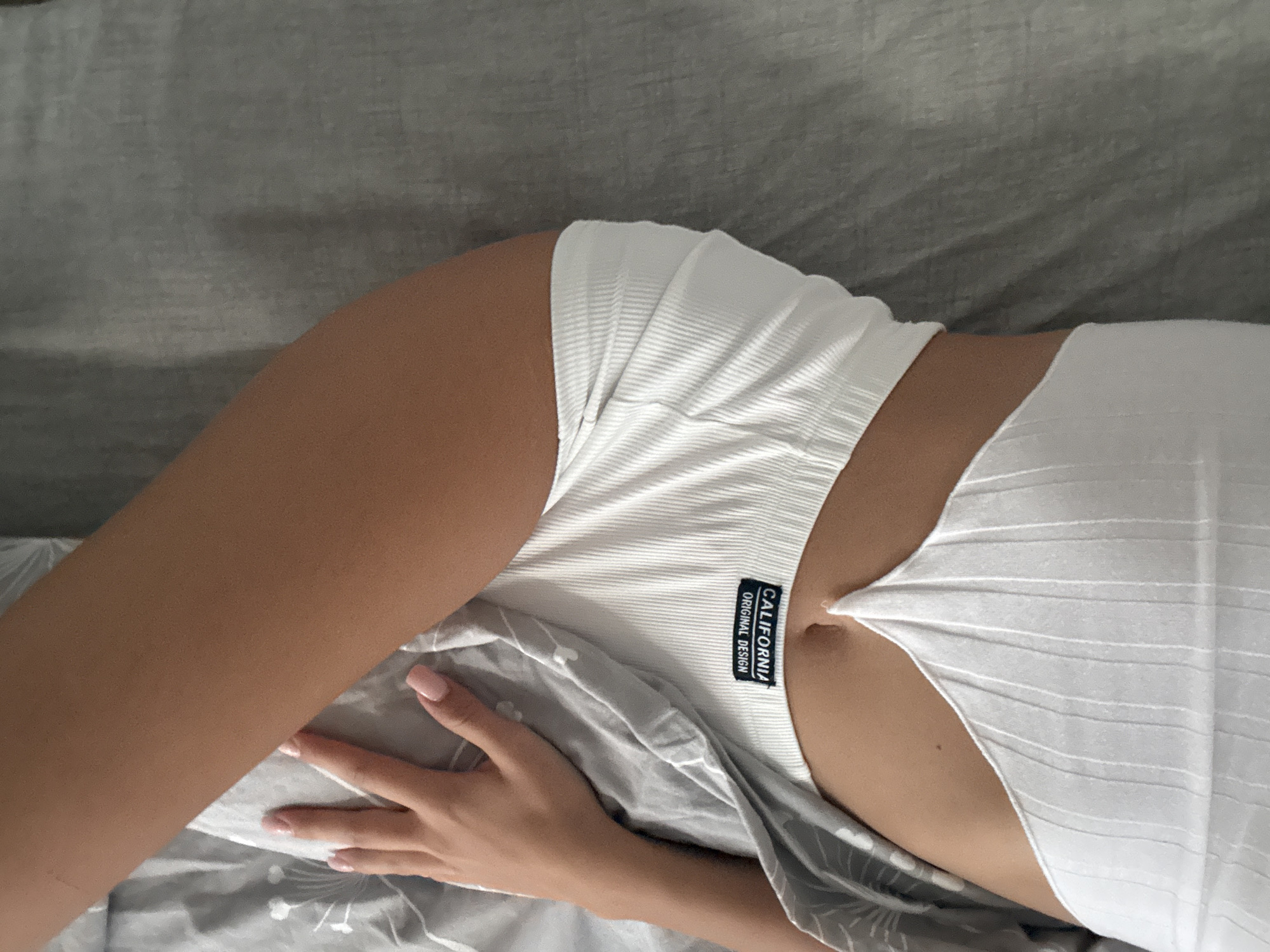 The image shows a close-up, overhead view of a person lying on a grey bed. The person's left arm and hand are visible, resting on a grey duvet with a subtle floral pattern. The person is wearing a white ribbed crop top with a black tag that reads "CALIFORNIA ORIGINAL DESIGN". The lower part of the crop top has a V-shaped cutout, revealing the person's midriff and navel. The background consists of the grey bedsheets, which have a textured appearance. The scene appears to be indoors, likely a bedroom. The location in Chișinău, Moldova, cannot be determined from the visual information provided in the image.