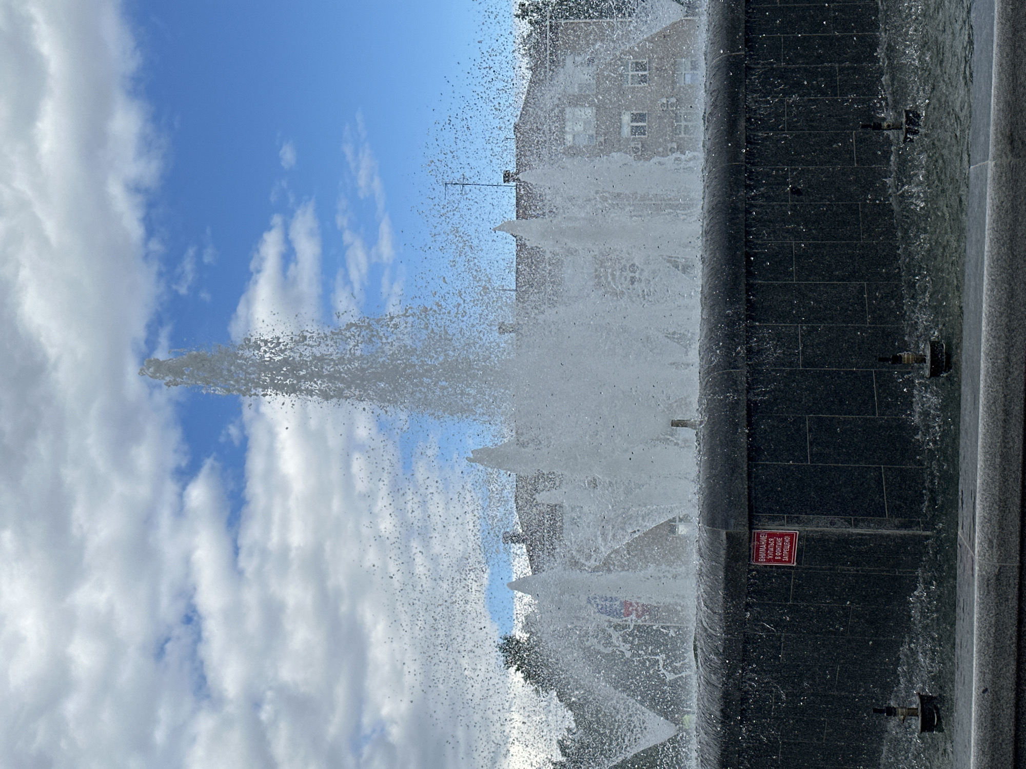 A fountain sprays water into the air under a cloudy blue sky. In the background, a building with windows and an antenna is visible, surrounded by trees. The fountain's dark, tiled basin has a red sign with Russian text that translates to "Attention! Swimming in the fountain is prohibited." Several water spouts are visible within the fountain, with water splashing around them. The foreground shows the edge of the fountain and the surrounding pavement.