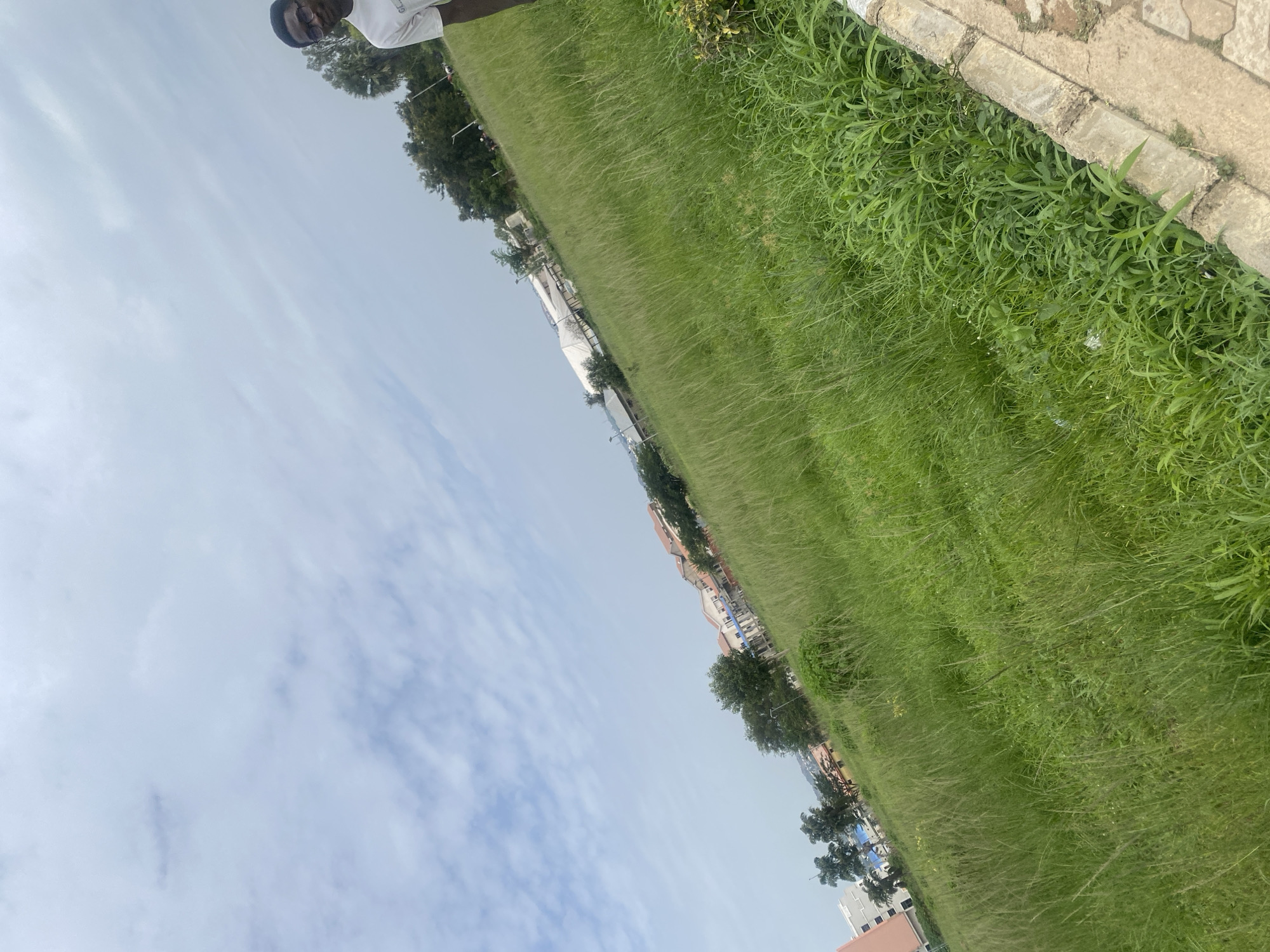 In Jos, Nigeria, a person is standing in a large, grassy field under a cloudy sky. The person, wearing a white t-shirt and glasses, is positioned in the right half of the frame, partially obscured. The field is characterized by tall, green grass, with some scattered bushes and a concrete curb running along the right edge of the image. In the background, buildings and trees are visible, suggesting a semi-urban or developing area. The overall scene depicts an outdoor environment with a focus on nature and a hint of ongoing construction or development.