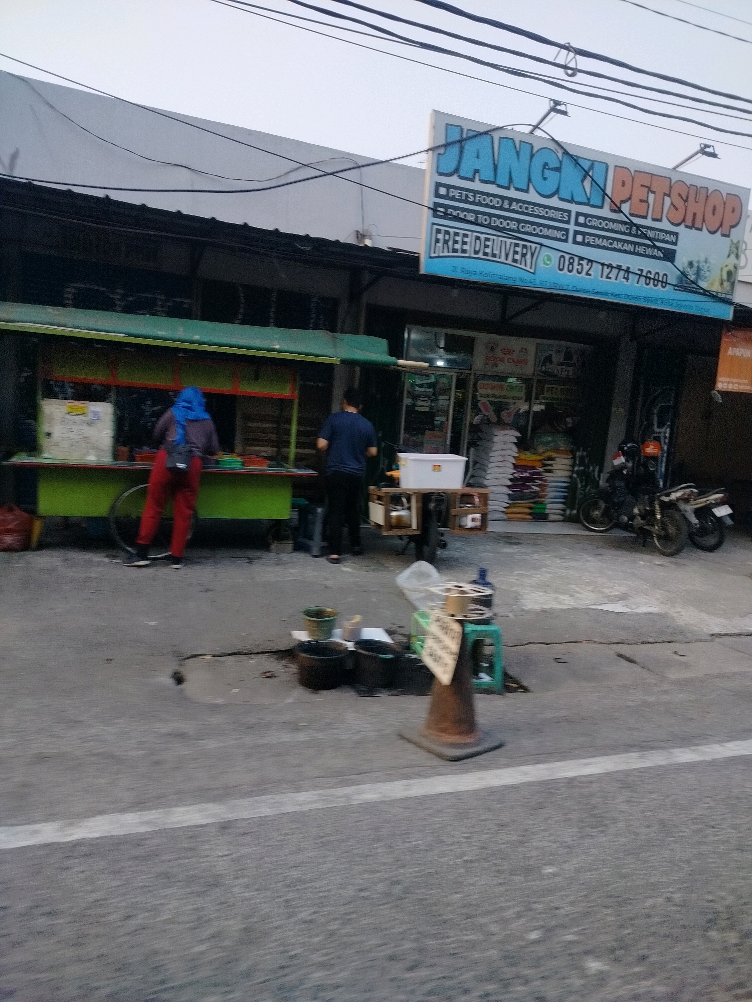 The image captures a street scene in East Jakarta, Indonesia, likely during daylight hours. Dominating the upper portion of the frame is a large, blue and orange sign that reads "JANGKI PETSHOP". Below this main text, smaller white lettering lists services offered: "PETS FOOD & ACCESSORIES", "GROOMING & NITIPAN", "DOOR TO DOOR GROOMING", and "PEMANAKAN HEWAN". A green phone number, "0852 1274 7600", is also visible, along with the text "FREE DELIVERY".
In front of the pet shop, a street food stall with a green awning is set up. Two individuals are present near the stall. One person, wearing a blue headscarf and red pants, appears to be working at the stall, with their back to the camera. Another person, wearing a dark blue t-shirt and black pants, stands facing the stall.
Several objects are scattered on the pavement in front of the businesses. A mobile cart with a white tub is positioned near the food stall. Motorcycles are parked in front of the pet shop. Further down the street, what appear to be sacks of pet food are stacked inside the shop. On the sidewalk in the foreground, several buckets and other containers are visible, along with a traffic cone and a small sign. The road surface in the foreground shows a white line, indicating a marked lane. The overall impression is a busy commercial street with a mix of businesses and street vendors.