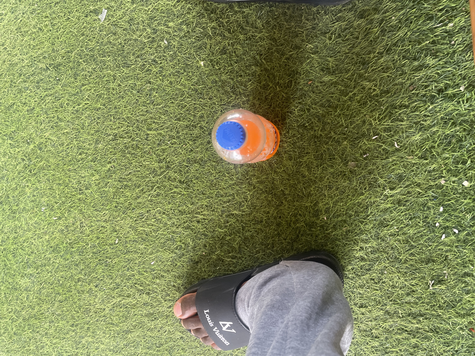 In Jauro Yinu, Nigeria, a close-up shot shows a person's left foot wearing a black Louis Vuitton slide sandal, positioned on artificial turf. Next to the foot, a partially filled orange beverage bottle with a blue cap rests on the same turf. The scene is observed from a high-angle perspective, looking down at the ground.