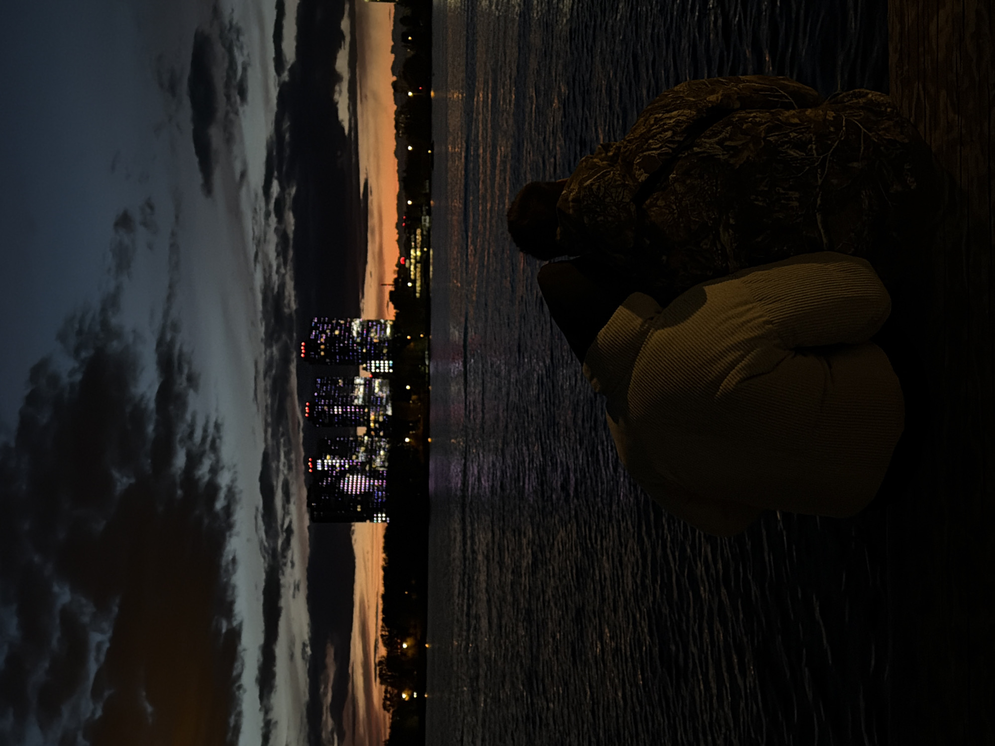 The image depicts a nighttime scene in Kazan, Russia, with a focus on a figure near a body of water.
On the left, the Kazan cityscape is visible under a cloudy sky, illuminated by city lights. The sky transitions from dark blue to orange near the horizon. The water reflects these lights, creating streaks of light on its surface.
To the right, a person sits near the water. The individual's head is resting on their knees. They are wearing what appears to be a light-colored corduroy jacket or sweater. Next to the person is a textured, dark-colored object, possibly a rock or a bag. The area around the person is dark.
The image does not reveal any specific details about the person's activities or interactions with the environment. The focus is on the juxtaposition of the urban landscape with the individual by the water.