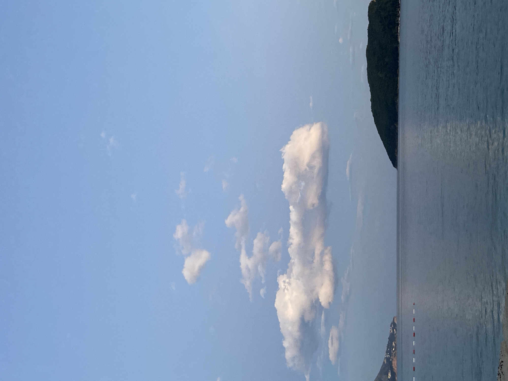 The image depicts a bright, sunlit coastal scene, likely near the city of Unknown. The view is oriented vertically, with a clear blue sky dominating the left side of the frame. Scattered cumulus clouds are present, some appearing elongated and vertical.
On the right side, the image transitions to a body of water, possibly a bay or the sea, which reflects the sky's color, although with a slightly darker, more muted tone. A dark, elevated landmass, perhaps a hill or headland covered in dense vegetation, is visible at the edge of the water. In the distance, near the horizon, is a series of red and white buoys marking a navigation channel. There is also a building or small settlement, though indistinct, visible near the water's edge.