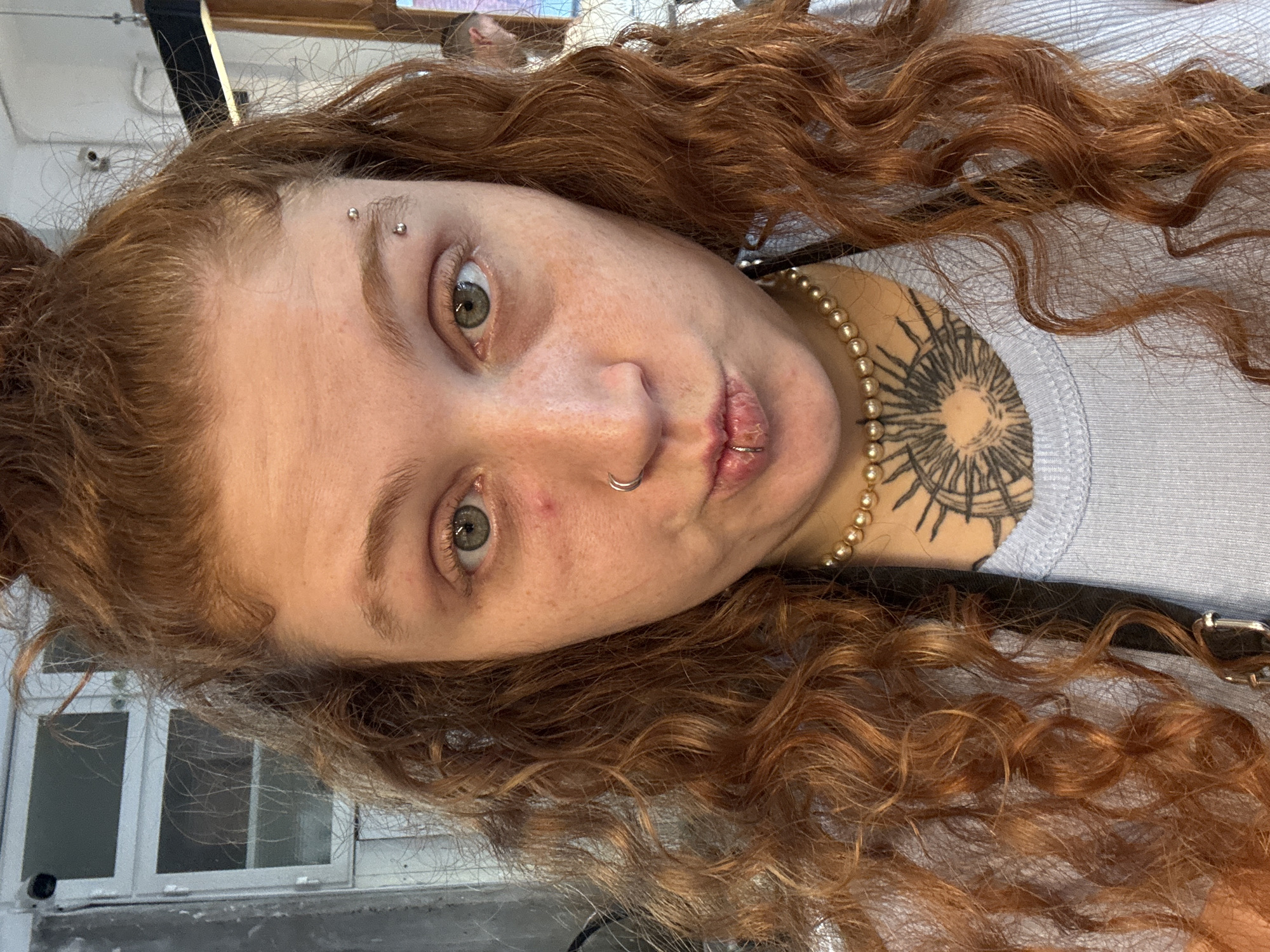A selfie of a red-haired individual with fair skin, freckles, and green eyes. The person has facial piercings on their eyebrow, nose, and lip. They wear a pearl necklace and have a tattoo on their upper chest or shoulder. The background includes a window with visible panes and a partially visible city environment that cannot be identified as Buenos Aires.