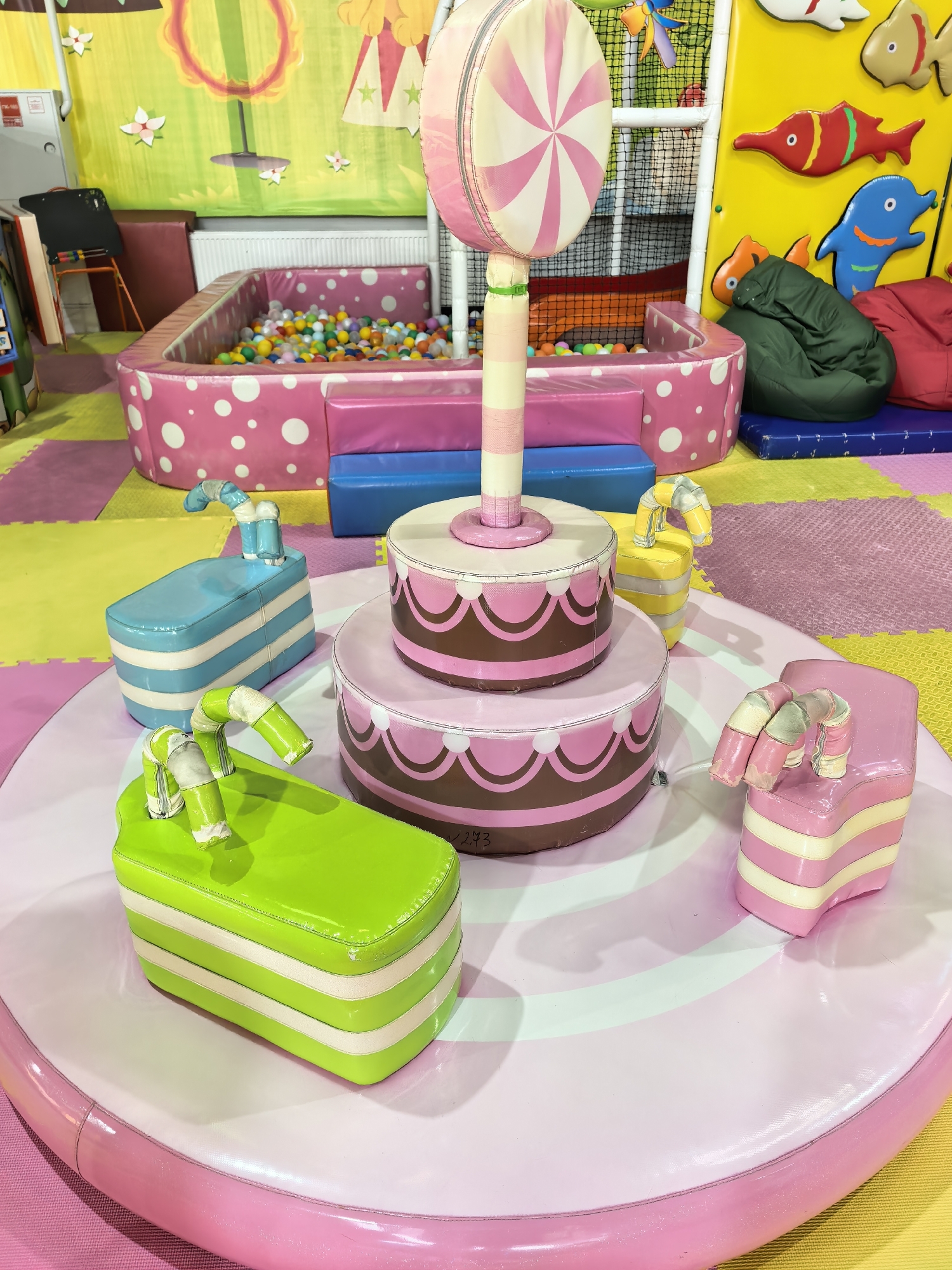 The image depicts a colorful indoor playground scene, likely located in Sterlitamak, Russia, featuring various play structures and decorations.
In the foreground, there is a circular platform adorned with pink and white stripes. Atop the platform, there is a multi-tiered cake-shaped structure with pink and brown frosting details, topped with a large lollipop prop. Surrounding the cake are four rectangular "chairs" or play structures in blue, green, yellow, and pink, each with a curved "straw" extending from them.
In the background, there is a ball pit enclosed by a pink border with white polka dots. Beyond the ball pit, there are padded walls and play equipment, including a yellow wall decorated with fish shapes and beanbag chairs.
The environment appears to be a commercial indoor playground, designed for children's entertainment. The overall impression is one of a playful and safe environment for kids to engage in imaginative play.