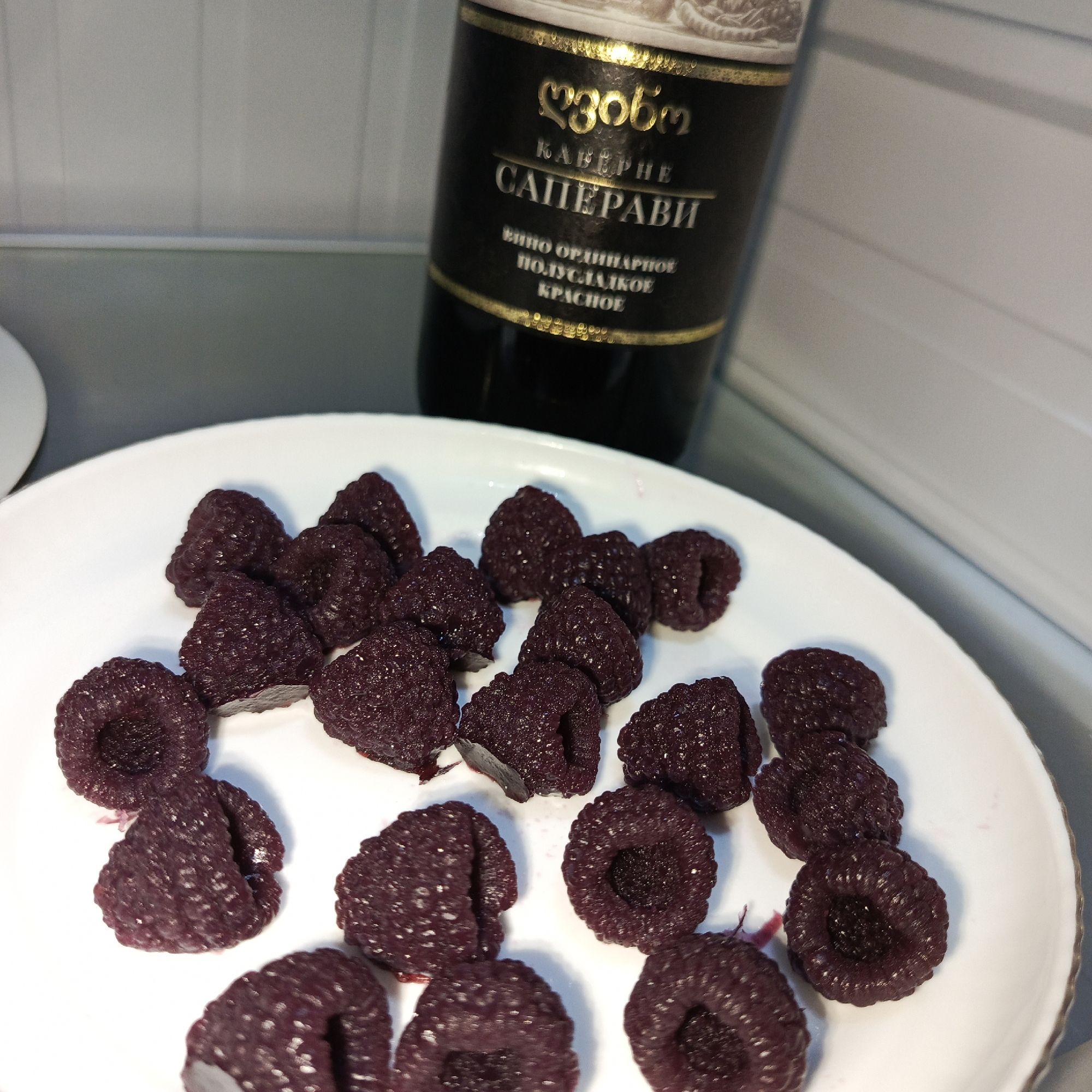 A static scene, likely located indoors in the city of Марковский, Russia, displays a white, slightly rimmed plate containing approximately 15-20 dark red to purplish raspberries. The raspberries appear moist and uniformly colored. Behind the plate, a dark glass wine bottle stands upright, partially visible. Its label is black with gold Cyrillic and Georgian lettering, identifying the contents as "КАБЕРНЕ САПЕРАВИ" (Cabernet Saperavi), an "ORDINARY SEMI-SWEET RED WINE." The background is a blurred light gray, suggestive of a refrigerator interior or a shelving unit. A small portion of a white object is visible on the far left edge of the frame. No individuals are present.