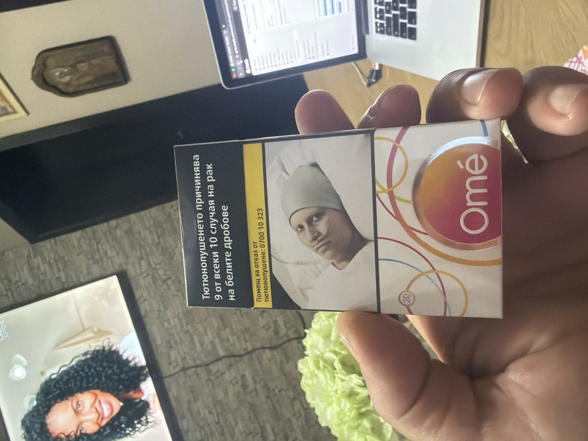 An overhead, slightly angled shot captures a person's hand holding a cigarette pack in what appears to be an indoor setting in Shumen, Bulgaria. The hand, with manicured nails, holds a white and colorful pack of "Omé" brand cigarettes.
The cigarette pack prominently features a graphic health warning in Bulgarian, taking up a significant portion of its surface. The text reads: "Тютюнопушенето причинява 9 от всеки 10 случая на рак на белите дробове" (Smoking causes 9 out of every 10 cases of lung cancer). Below this, another warning states: "Помощ за отказ от тютюнопушене: 0700 10 323" (Help to quit smoking: 0700 10 323). The image accompanying the warning shows a somber-looking individual, possibly a child or young adult, wearing a medical head covering and a nasal cannula, indicative of illness. The number "20" is visible on the pack, likely indicating the quantity of cigarettes.
In the upper background, a silver laptop, resembling a MacBook, is open and displaying content on its screen. A charging cable or power cord is visible nearby. To the lower left, another screen or tablet displays a close-up image of a smiling woman with dark curly hair. In the lower right foreground, a lush green, possibly artificial, floral arrangement adds a touch of color. Further back in the upper left, a small, dark rectangular object, possibly a religious icon or plaque, is mounted on a light-colored wall or shelf. The overall lighting suggests a daytime setting.