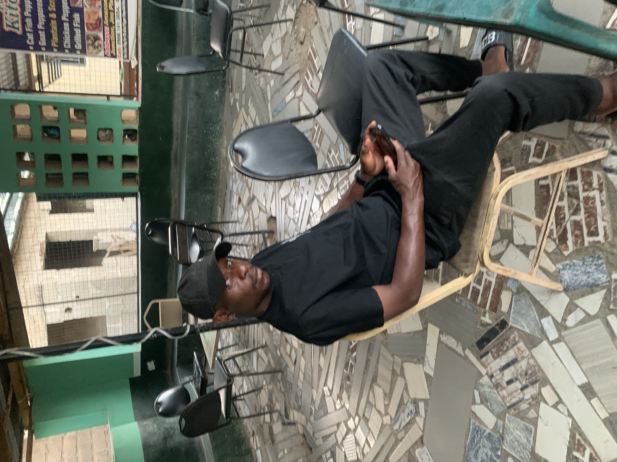The image depicts a man sitting in a chair in an outdoor setting. He is wearing a black t-shirt, black pants, and a black baseball cap. He is holding a mobile phone. The ground is covered in mosaic tiles. Surrounding him are several empty chairs. In the background, there is a building and a sign that advertises food items like "Cat Fish Pepper", "Goat Peppered" and "Chicken Pepper". The sign also includes phone numbers. The location is implied to be New Stadium, Nigeria, based on the text on the sign.