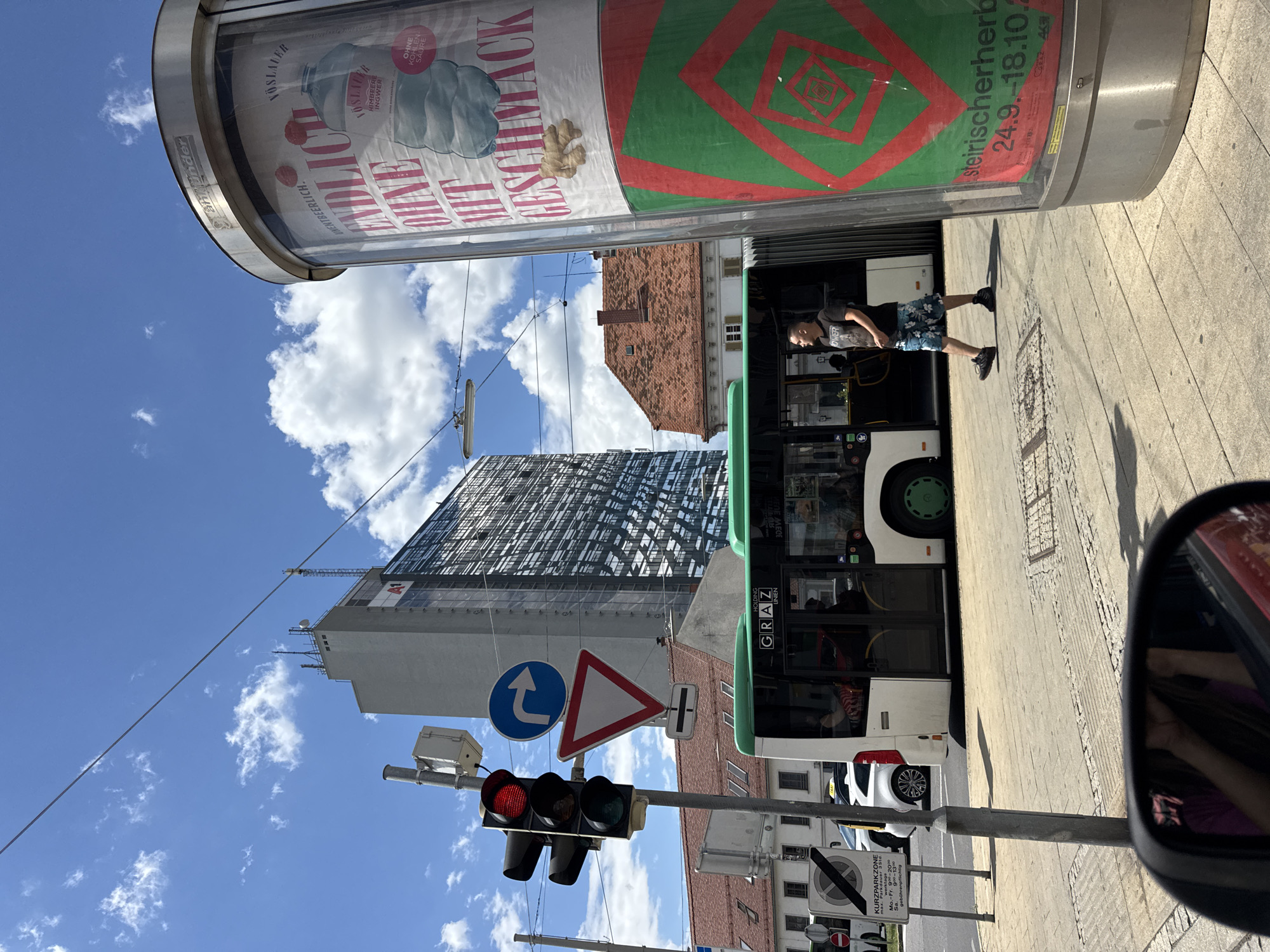 A street scene in Graz, Austria, features a modern building with a glass facade, a bus with "GRAZ UNIEN" written on its side, a man walking, and a large cylindrical advertising pillar. The sky is blue with scattered clouds. Traffic lights are red and green. A yield sign and a turn right sign are visible. A parked white car and a sign indicating "KURZPARKZONE" are also present. The advertising pillar displays a large poster for "VÖSLAUER" water, with text in German. The date range "24.9. - 18.10." is also visible on the poster. A partial view of a car's rearview mirror shows a person's reflection.