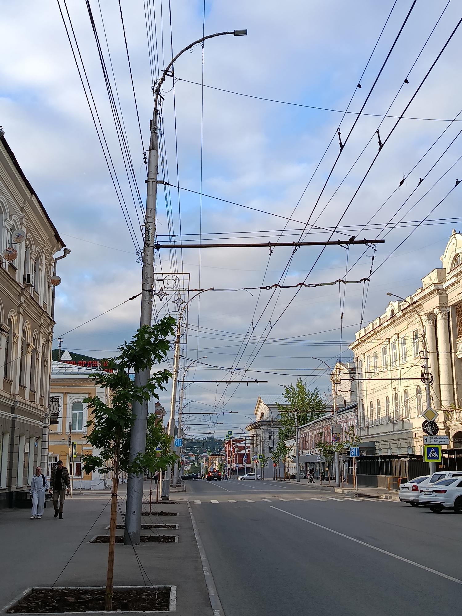 This is a street-level, eye-level, medium-shot photograph taken in Saratov, Russia, likely on or near Moskovskaya Street, as indicated by signage. The scene depicts a city street with prominent overhead wires, characteristic of a trolleybus system.
On the left side of the street, a building with ornate, light-colored architecture and arched windows is visible. A narrow sidewalk runs alongside this building, where two individuals, a man and a woman, are walking away from the camera. The man is wearing a patterned jacket and dark pants, while the woman is dressed in a white tracksuit. Young trees in planters are spaced along the sidewalk.
The street itself is paved, with white lane markings. Several cars are present, including two white sedans parked on the right side of the road. The right side of the street features buildings with a similar classical architectural style, with columns and numerous windows. Traffic signals, streetlights, and signage, including a yellow diamond-shaped sign with a black arrow and text in Russian, are also visible.
The sky is partly cloudy, with a mix of blue sky and white clouds. The overall impression is an urban environment during daylight hours, with the complex network of overhead wires dominating the upper portion of the image.