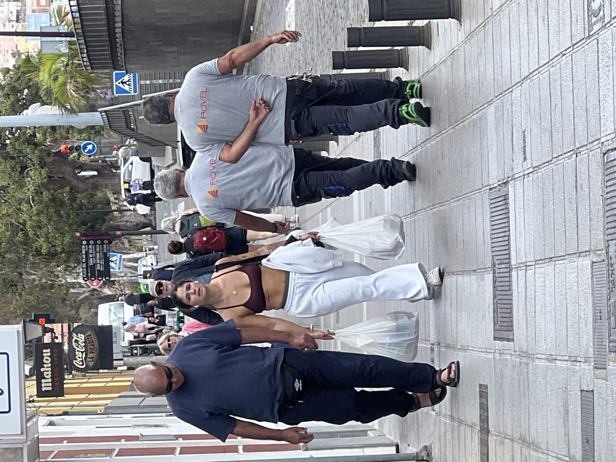 The image shows a street scene in Las Palmas De Gran Canaria, Spain, with several people walking on a sidewalk. In the foreground, an older man wearing a navy blue shirt, dark pants, and sandals walks toward the camera carrying a white plastic bag. Behind him, a young woman in a burgundy sports bra and white pants also carries a white plastic bag. Further back, a group of people is walking in the same direction. In the top of the image, two men wearing grey "ROVEL" branded t-shirts and dark work pants are walking away from the camera. Street signs and advertisements for "Mahou" and "Coca-Cola" are visible on the left side of the frame. A palm tree and various buildings are in the background. The street appears to be paved with grey tiles.