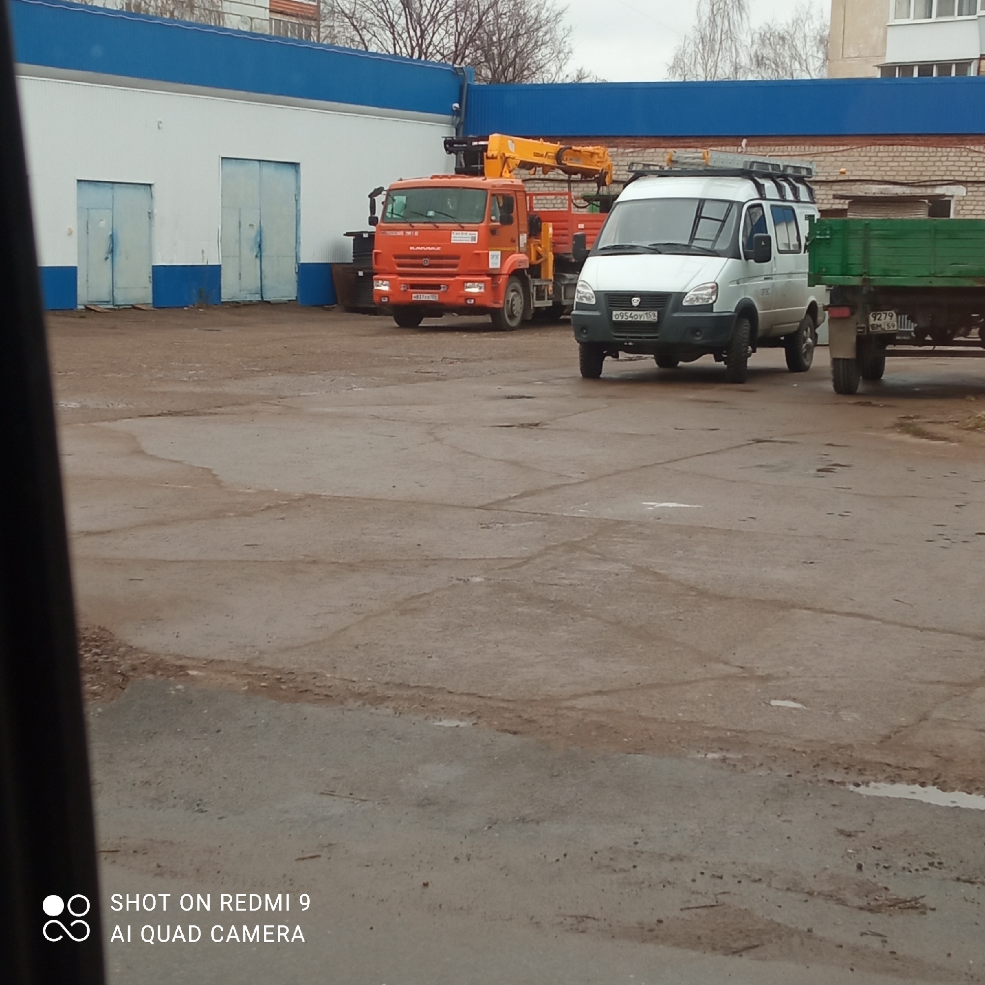 The image shows an exterior shot of what appears to be a paved service yard in Chaikovsky, Russia. In the background is a one-story building with a white facade, a blue roof, and blue accents around the base. Two large service doors are visible on the building.
Three vehicles are present in the midground. An orange "KAMAZ" truck is parked towards the left, and a white van with a roof rack is parked beside it. Connected to the white van is a small green trailer.
The ground is paved and appears to be weathered, with cracks and some patches of discoloration, and a wet surface.
The sky is overcast, suggesting it may be a cloudy day. The photo appears to have been taken from inside a vehicle, as part of the window is visible on the left side of the frame. The text at the bottom indicates the photo was taken with a Redmi 9 AI Quad Camera.