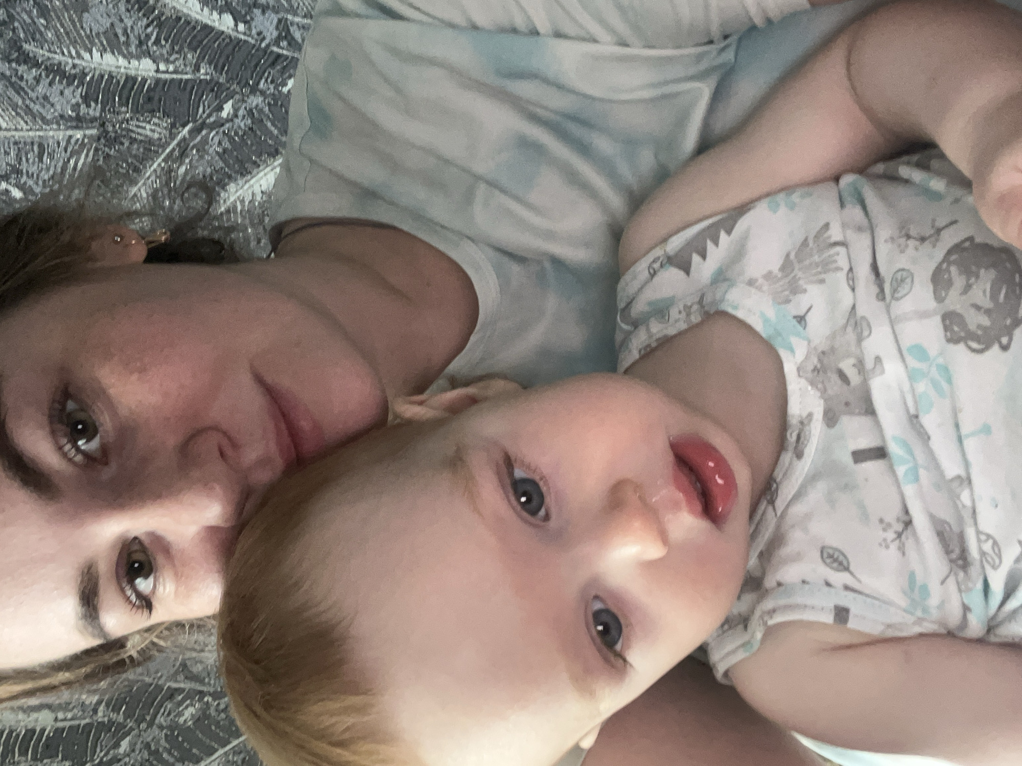 The image depicts an adult female and an infant in an indoor setting within the city of Chaikovsky, Russia. The adult female, positioned in the upper frame, has light brown hair, brown eyes, and fair skin, wearing a light blue and white mottled t-shirt and small gold-colored hoop earrings. She looks directly into the camera. An infant, with reddish-blonde hair and blue eyes, is situated in front of the adult, resting against her. The infant has a small amount of clear liquid on the upper lip and below the nose and is dressed in a white and light blue sleeveless top featuring a pattern of stylized trees, leaves, and various animal figures. The background consists of a wall covered with dark grey or black wallpaper patterned with light-colored, stylized leaves.