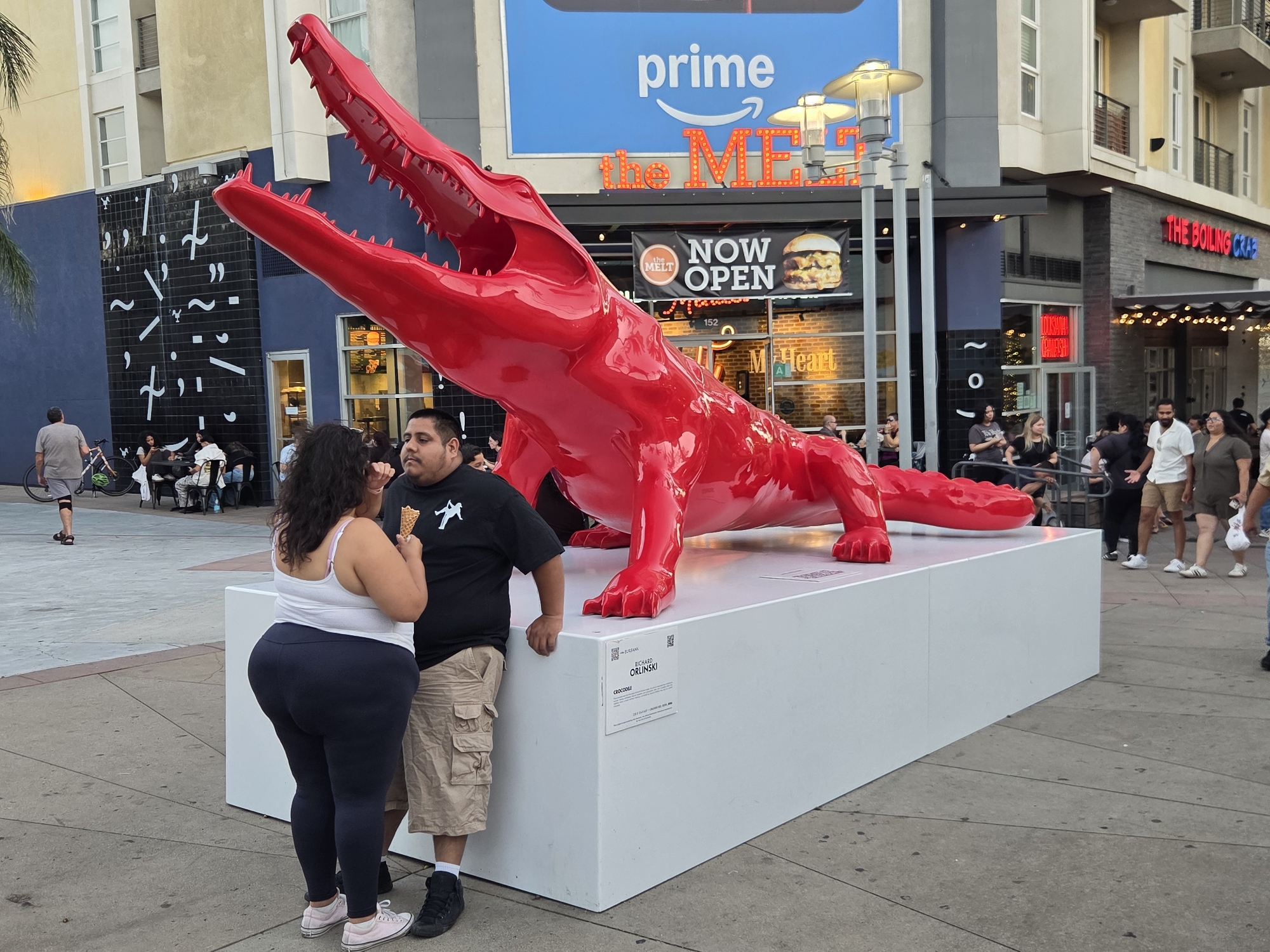 A life-sized, vibrant red crocodile sculpture, identified by a plaque on its white pedestal as "CROCODILE" by Richard Orlinski, is prominently displayed outdoors. The sculpture's mouth is open, revealing sharp teeth. Two individuals, a man and a woman, stand next to the pedestal, appearing to interact with each other. The man, wearing a black t-shirt and khaki shorts, holds an ice cream cone. The woman, in a white tank top and dark leggings, is also holding an ice cream cone. The background reveals a commercial area with storefronts, including a restaurant with a "NOW OPEN" banner featuring a burger, and a larger sign that reads "prime the MELT". The overall setting appears to be a public urban space, possibly part of a shopping or entertainment district, consistent with the city of Burbank, California, United States of America. Other people are visible in the background, suggesting a lively public area.