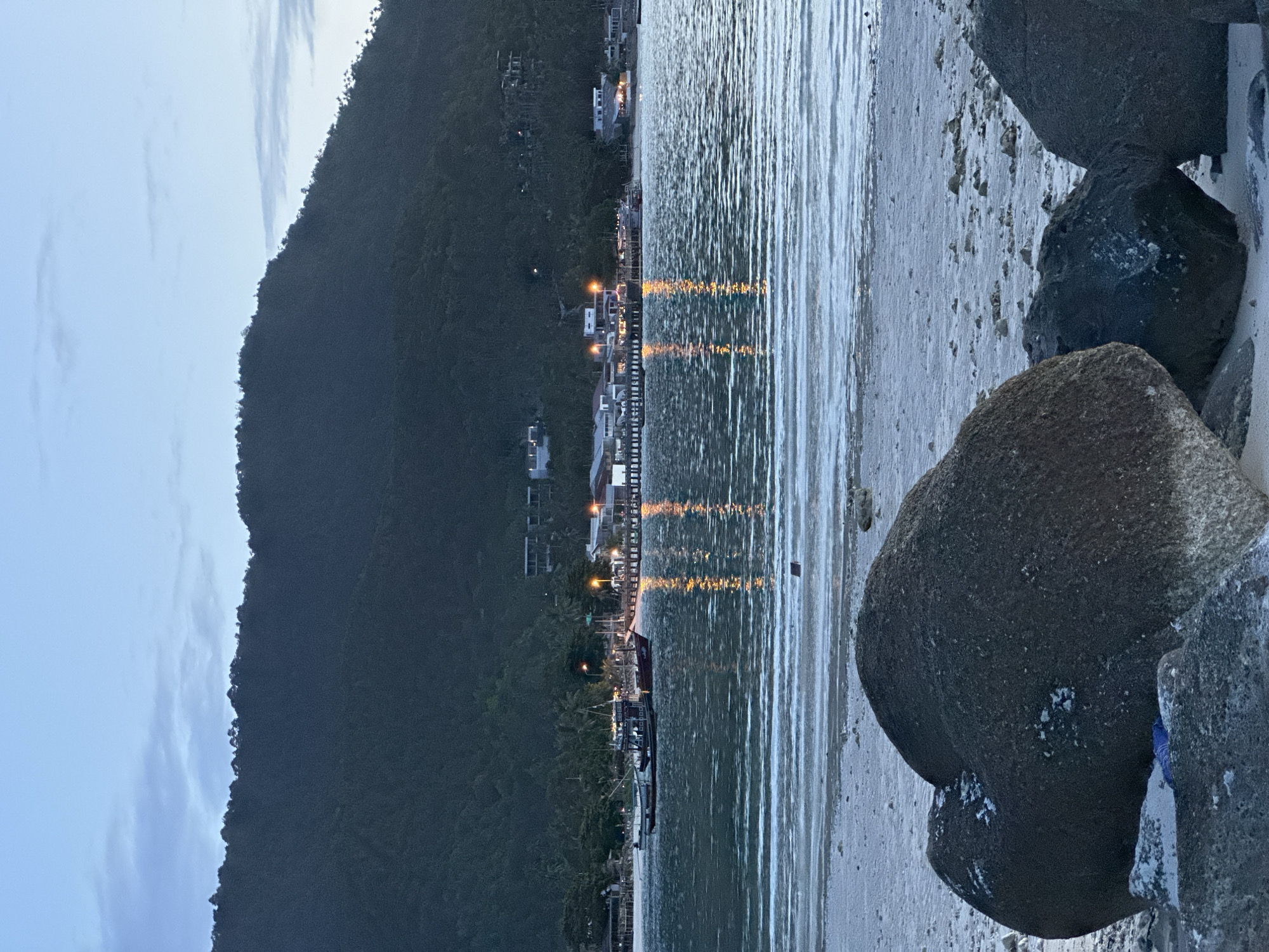 The image depicts a twilight scene at a coastal area in Tambon Ko Phangan, Thailand. In the foreground, large dark rocks sit on a sandy beach. Beyond the beach, a body of water, likely the sea, is visible with small waves creating a textured surface. A wooden pier extends into the water, lined with buildings and illuminated by artificial lights, casting reflections on the water's surface. Several boats are moored near the pier. In the background, a densely forested hill rises steeply, with scattered buildings visible along its lower slopes and near the shoreline. The sky above is a pale blue with faint clouds, indicating the transition from day to night. The overall impression is a serene, inhabited coastal landscape.