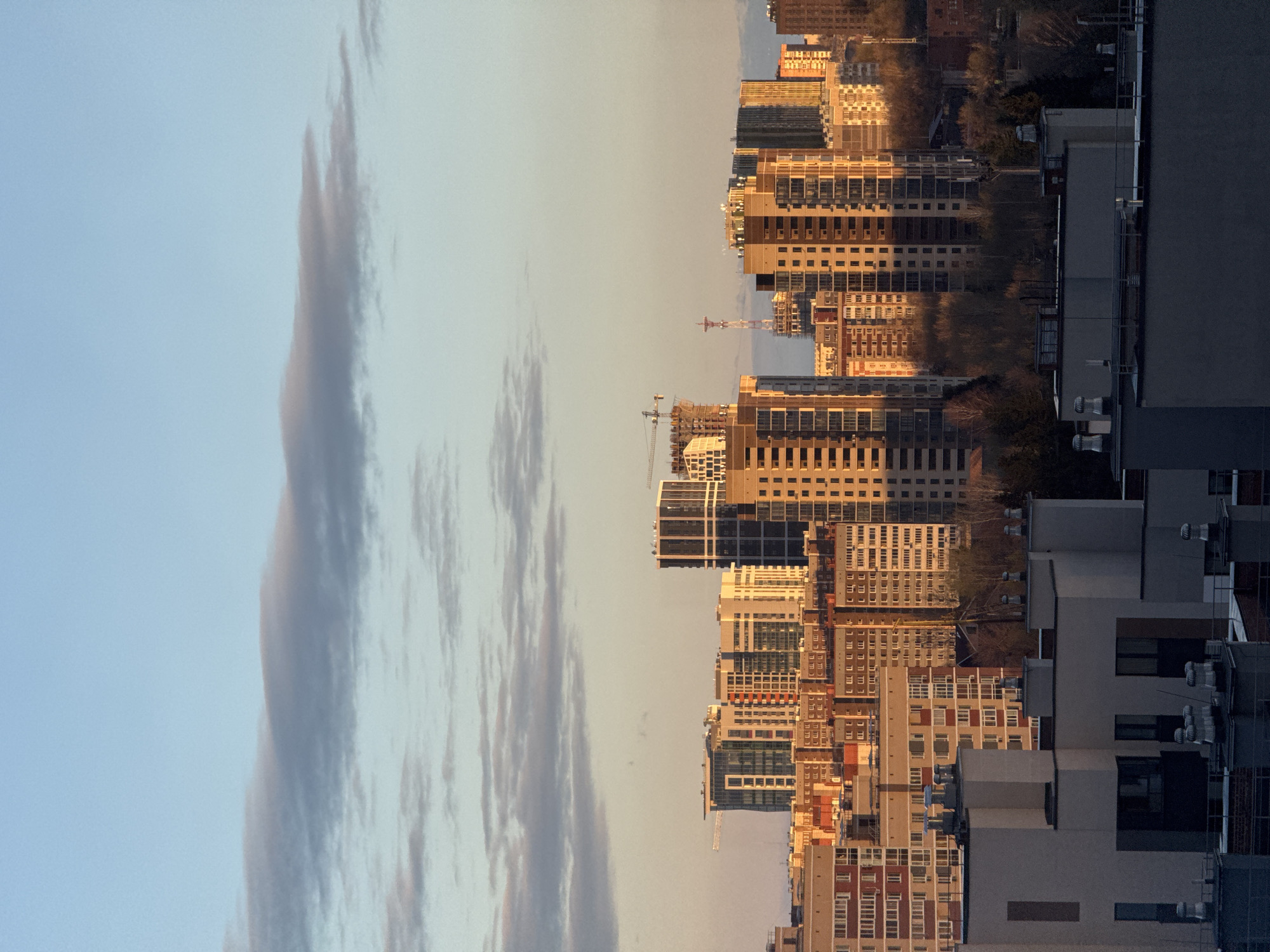 The image captures a modern cityscape, likely Izhevsk, Russia, under a clear sky, bathed in the warm, low-angle light of either sunrise or sunset. The sky, occupying the upper portion of the frame, transitions from a soft blue at the top to a warm, pale yellow near the horizon. Several long, wispy clouds streak across the sky, subtly illuminated by the golden light.
The lower two-thirds of the frame are dominated by a dense cluster of high-rise buildings, predominantly modern in architectural style, featuring numerous windows and balconies. Many of these structures, particularly in the mid-ground, are brightly lit by the golden hour sunlight, emphasizing their facades and patterns. Buildings closer to the foreground and on the far right appear darker, either facing away from the sun or cast in shadow by other structures.
Visible within the urban sprawl are at least two prominent tower cranes, signifying ongoing construction and development within the city. Patches of dark ground and scattered trees are discernible between the buildings, suggesting small green spaces or undeveloped plots amidst the dense concrete landscape. The scene is static, portraying the city in a serene moment of natural light. No visible text is discernible in the image.