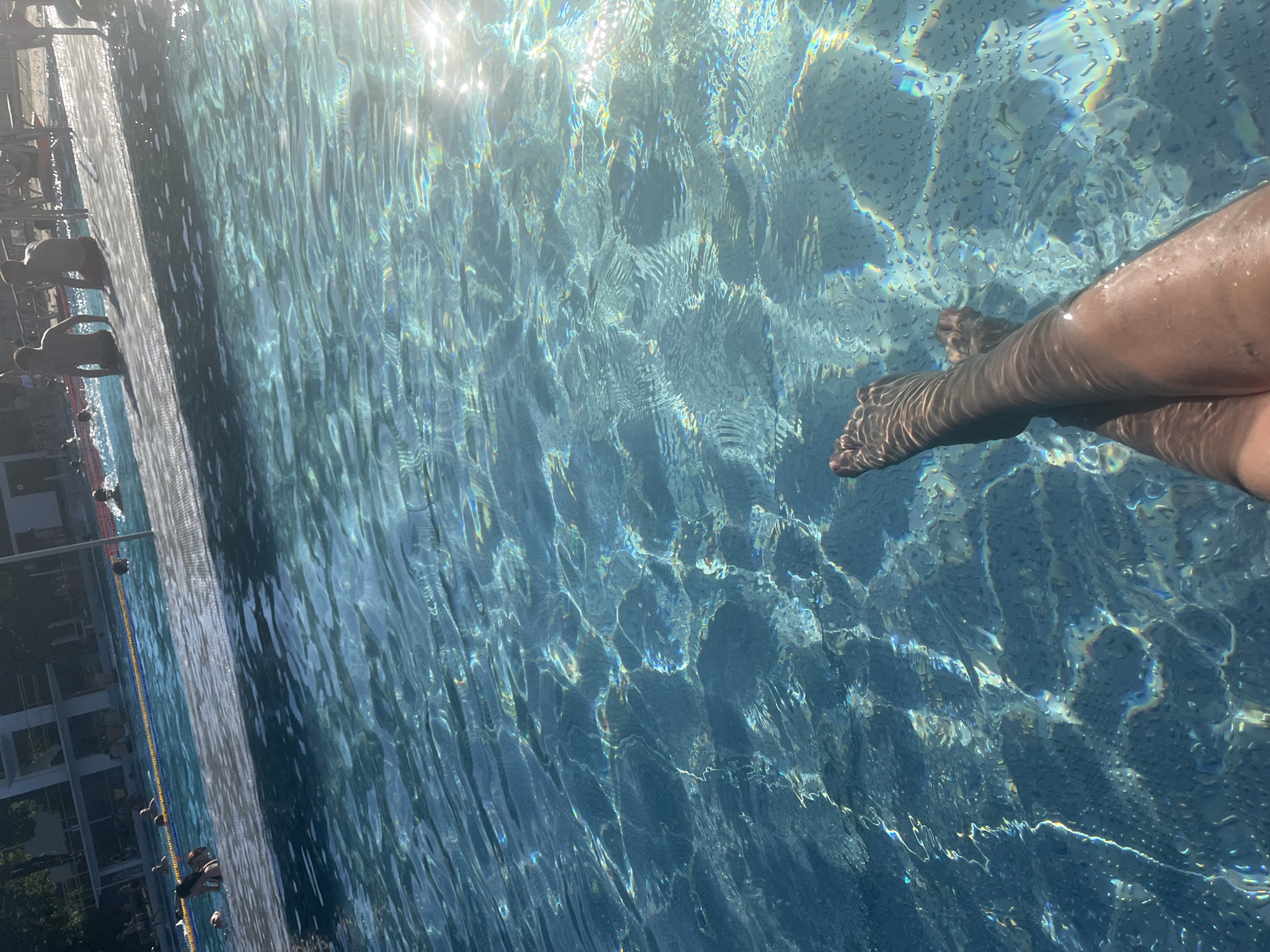 The image shows a person's legs and feet submerged in clear blue water of a swimming pool. The sunlight glints off the water's surface, creating a shimmering effect. In the background, there are other people in the pool and on the poolside, as well as some trees and a building. The setting appears to be an outdoor swimming pool on a sunny day.