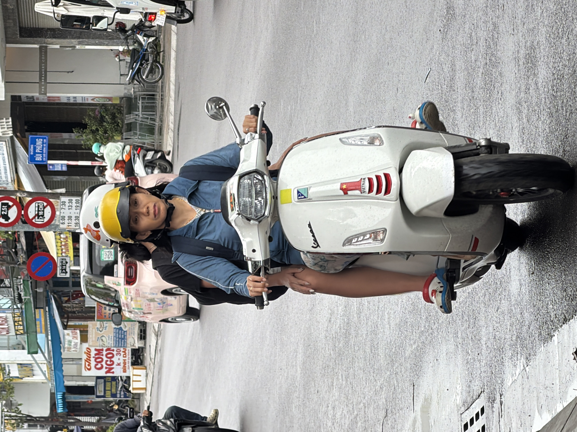 In Nha Trang, Vietnam, a woman wearing a yellow helmet and blue jacket rides a white Vespa scooter. She has dark hair and is looking towards the camera. A passenger with a white and orange helmet is seated behind her, also facing forward. Both individuals appear to be wearing casual clothing. The scooter has "Vespa" branding and a sticker with a red "1" and "VIETNAM" on the front. The road surface is wet, suggesting recent rain. Other vehicles, including motorcycles and a pink car adorned with stickers, are visible in the background, along with shops and street signs. One sign indicates "BUI PHUNG" street. The scene captures a common mode of transportation in the urban environment of Nha Trang.