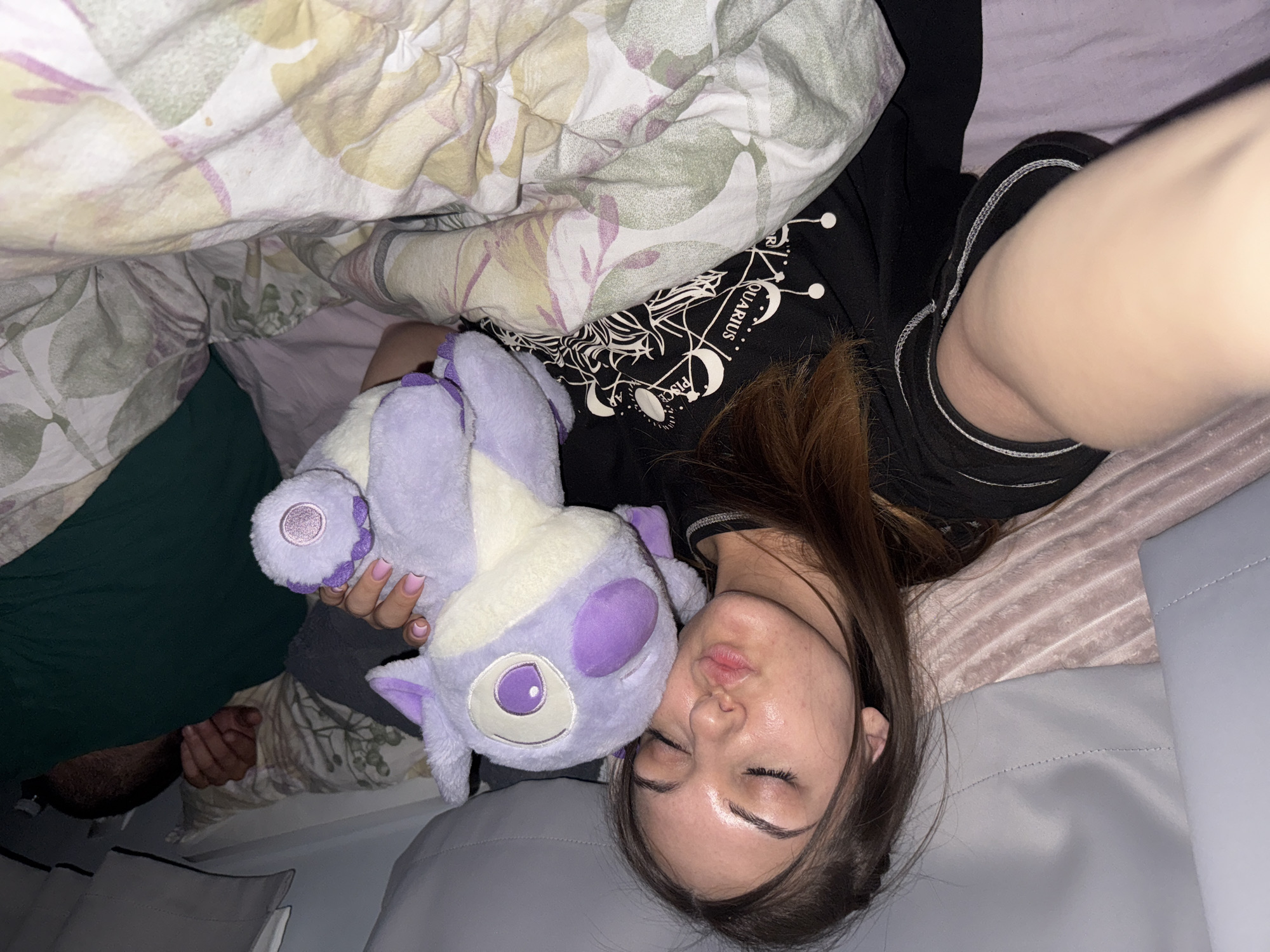 A young woman lies in bed, holding a purple plush toy. The bed is covered with patterned bedding. The woman wears a black shirt with a zodiac design. Her eyes are closed. No indication shows that the location is Sofia, Bulgaria.