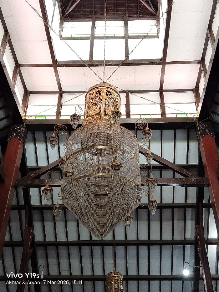 The image shows a large, ornate chandelier hanging from the ceiling of a room. The chandelier is made of gold and crystal and is suspended by chains. The room has a wooden ceiling with exposed beams. The walls are white and the ceiling has a white painted surface. There are several smaller chandeliers visible in the image. The image was taken in a building located at latitude -6.327, longitude 108.322 in Indramayu, Indonesia.