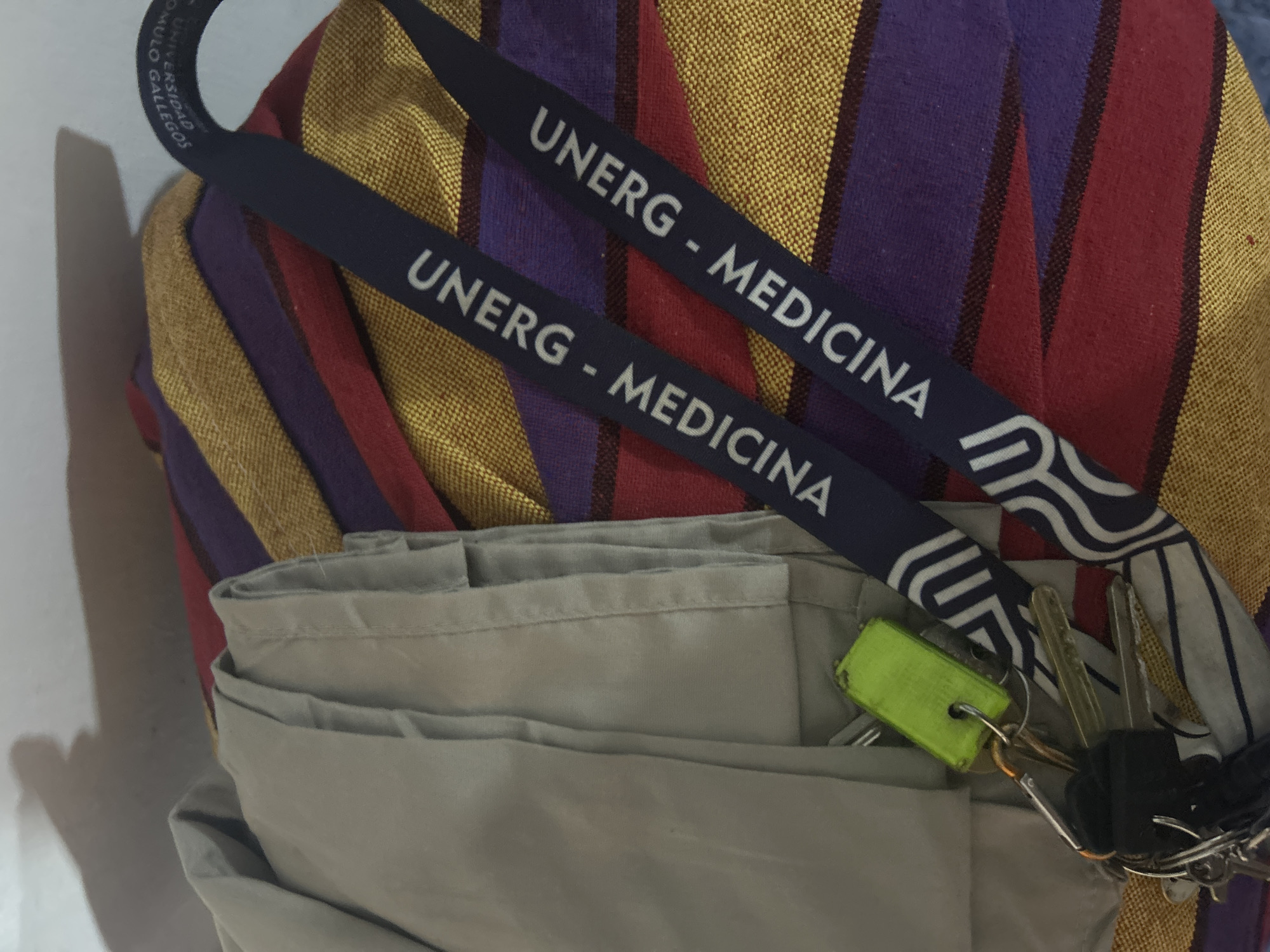 The image shows a close-up of several items. Two dark blue lanyards with white lettering reading "UNERG - MEDICINA" are prominent. The lanyards appear to be hanging from a striped textile of gold, purple, and red. Below the lanyards and the textile is a folded, light tan garment. A set of keys with a neon green tag is draped on the garment. The keys are various shapes and sizes. The image was likely taken indoors. There is no indication of the location in the city of San Juan de Los Morros, Venezuela.
