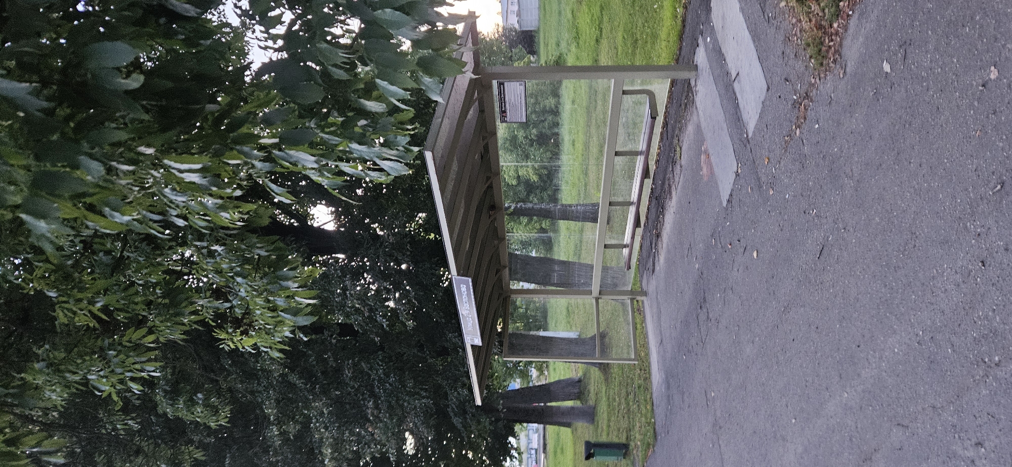 The image shows a bus stop in Vitsebsk, Belarus. The bus stop is located on a paved area next to a grassy lawn. There are several trees in the background, and the sky is overcast. The bus stop itself is made of metal and glass, and it has a bench for passengers to sit on. There is also a trash can next to the bus stop. In the distance, there are some buildings visible. The overall scene is peaceful and quiet, suggesting a normal day in a suburban area.