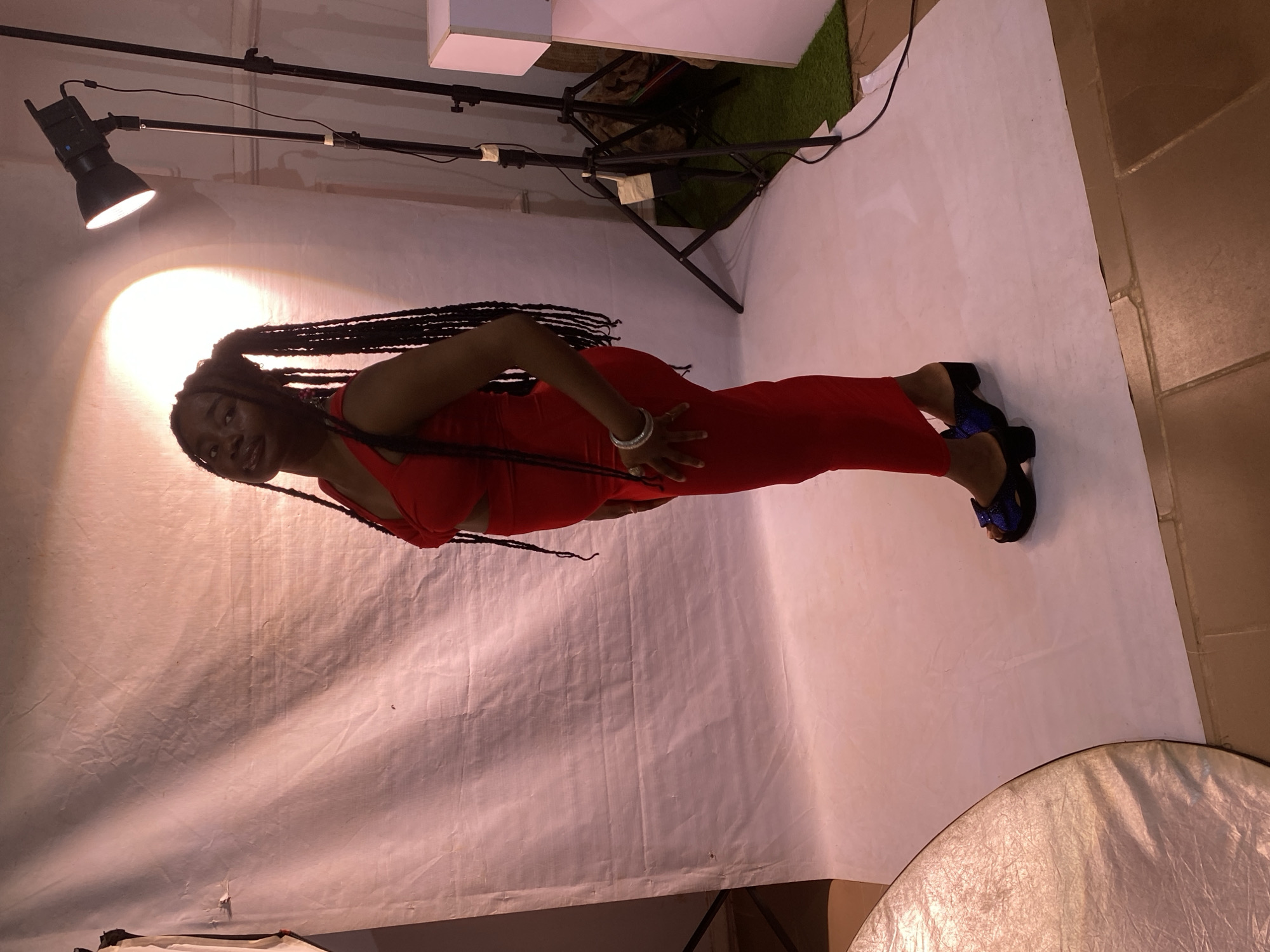 The image shows a young woman with long braided hair wearing a red jumpsuit and black high-heeled shoes with blue bows. She is standing in a studio setting with a white backdrop. There are studio lights and equipment visible in the background, including a large softbox on the right side of the frame. The lighting creates shadows on the backdrop. The woman is looking at the camera and smiling. She is wearing a silver bracelet on her left wrist. The scene appears to be professionally lit, suggesting a photoshoot or a similar event. There is no visible text in the image. The setting suggests an indoor location, possibly a photography studio. The overall mood is bright and positive.