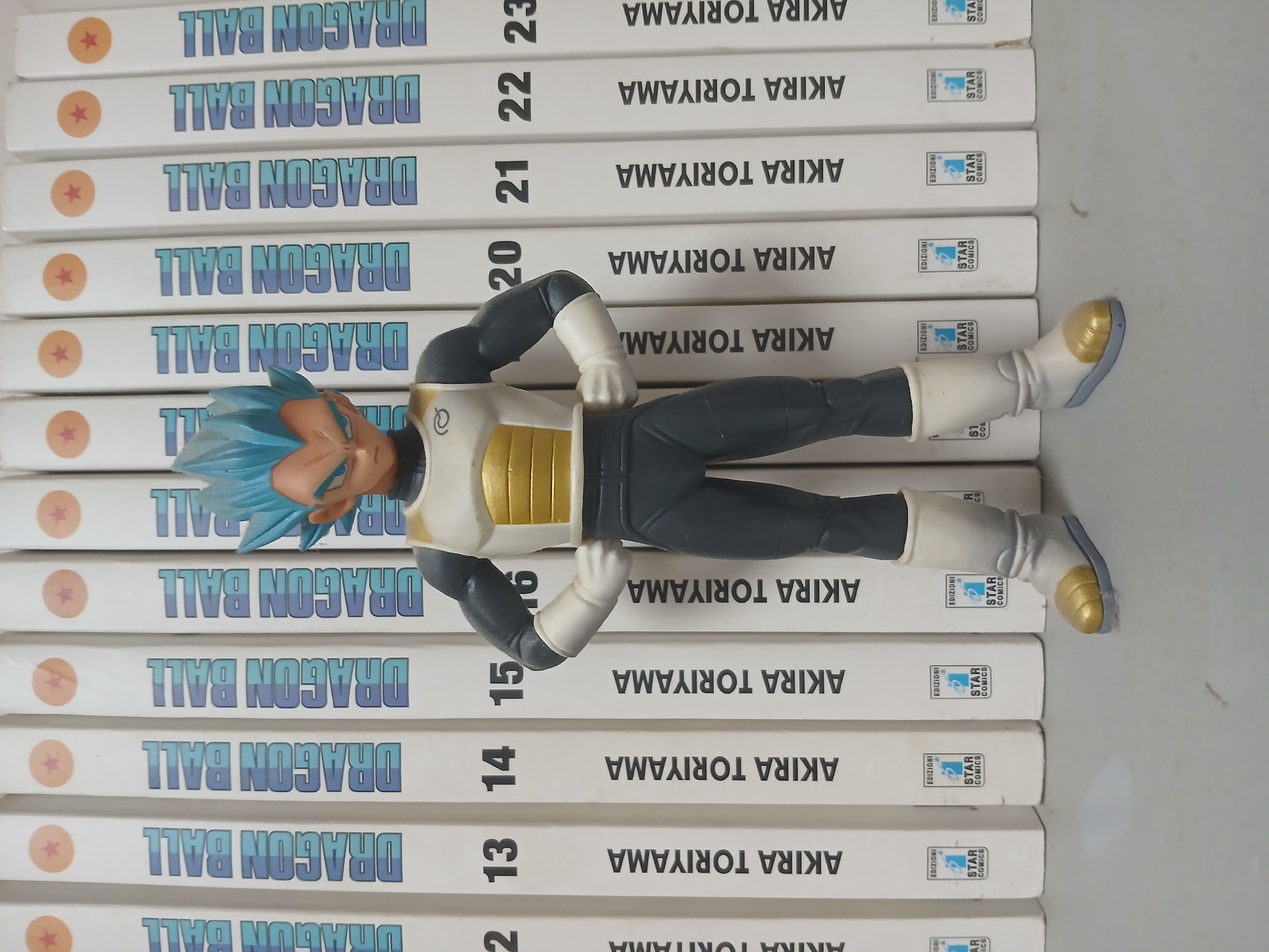 A male figure, resembling the character Vegeta from Dragon Ball, stands centered in the foreground. The figure is wearing a white and gold chest plate with gray sleeves and pants, and white boots with gold accents. Behind the figure, a row of vertically oriented books or manga volumes are arranged. The spines of these volumes are predominantly white with the text "DRAGON BALL" and "AKIRA TORIYAMA" in a blue font. Numbers such as "13", "14", "15", "16", "20", "21", "22", and "23" are visible on some of the spines. The bottom portion of the book spines show logos with the text "EDIZIONI STAR COMICS". The background appears to be a plain, light-colored surface. The overall scene is indoors, likely on a shelf or table. The image composition suggests a product display or collection.
