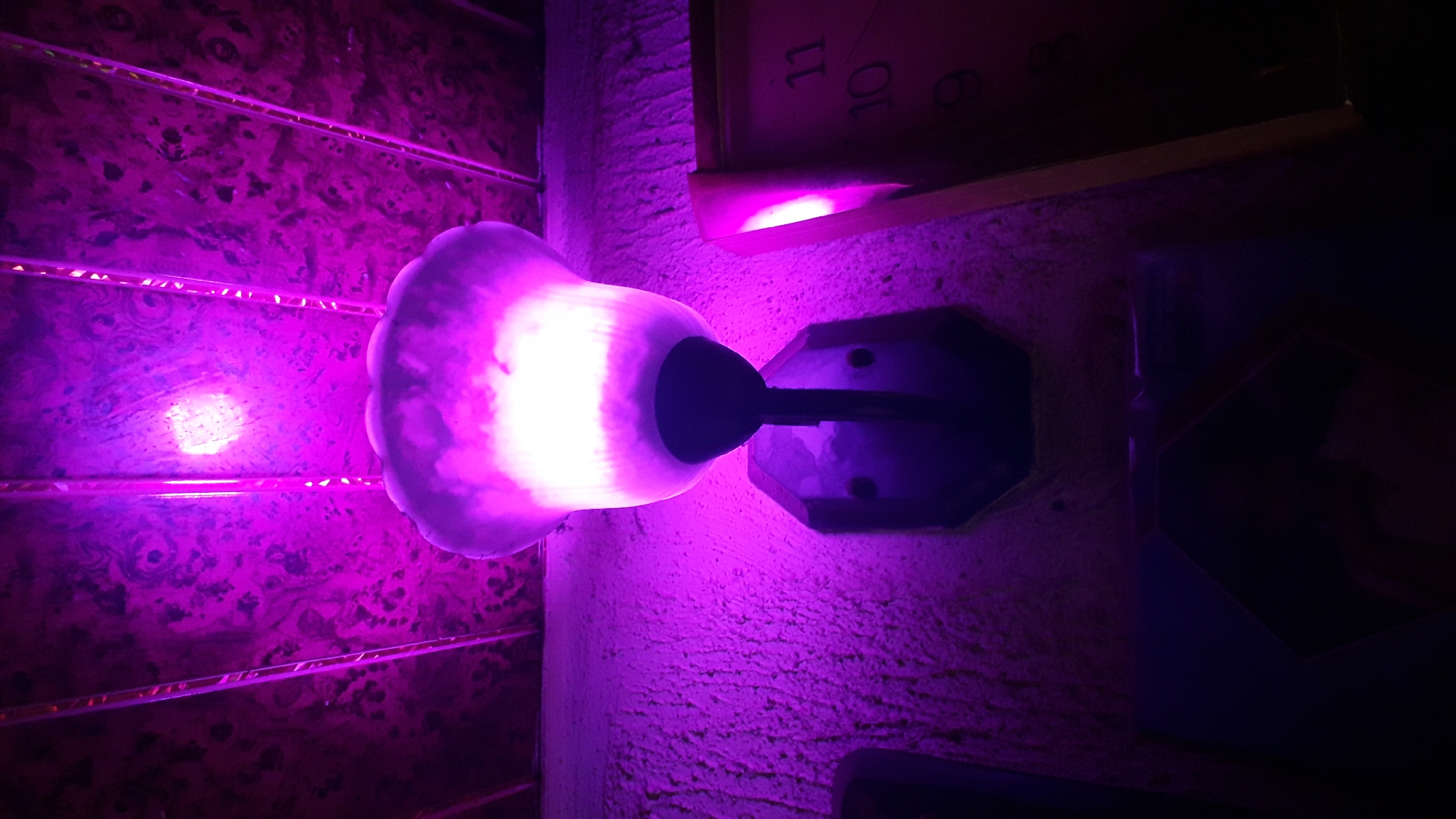 This is a low-angle, upward shot of a purple-lit wall sconce mounted on a textured wall. The sconce features a bell-shaped frosted glass shade with a dark central element, illuminated by a bright white light emanating from within. Above the sconce, the ceiling is paneled with a dark, swirling wood-grain pattern, also illuminated with purple light. To the right of the sconce, a portion of a clock face with red numbering is visible, displaying the numerals 9, 10, and 11. A thin, vertical wooden frame borders the clock face. The overall ambiance is dimly lit with strong purple and white illumination, creating a dramatic and somewhat eerie atmosphere. The location is identified as Nkpor, Nigeria.