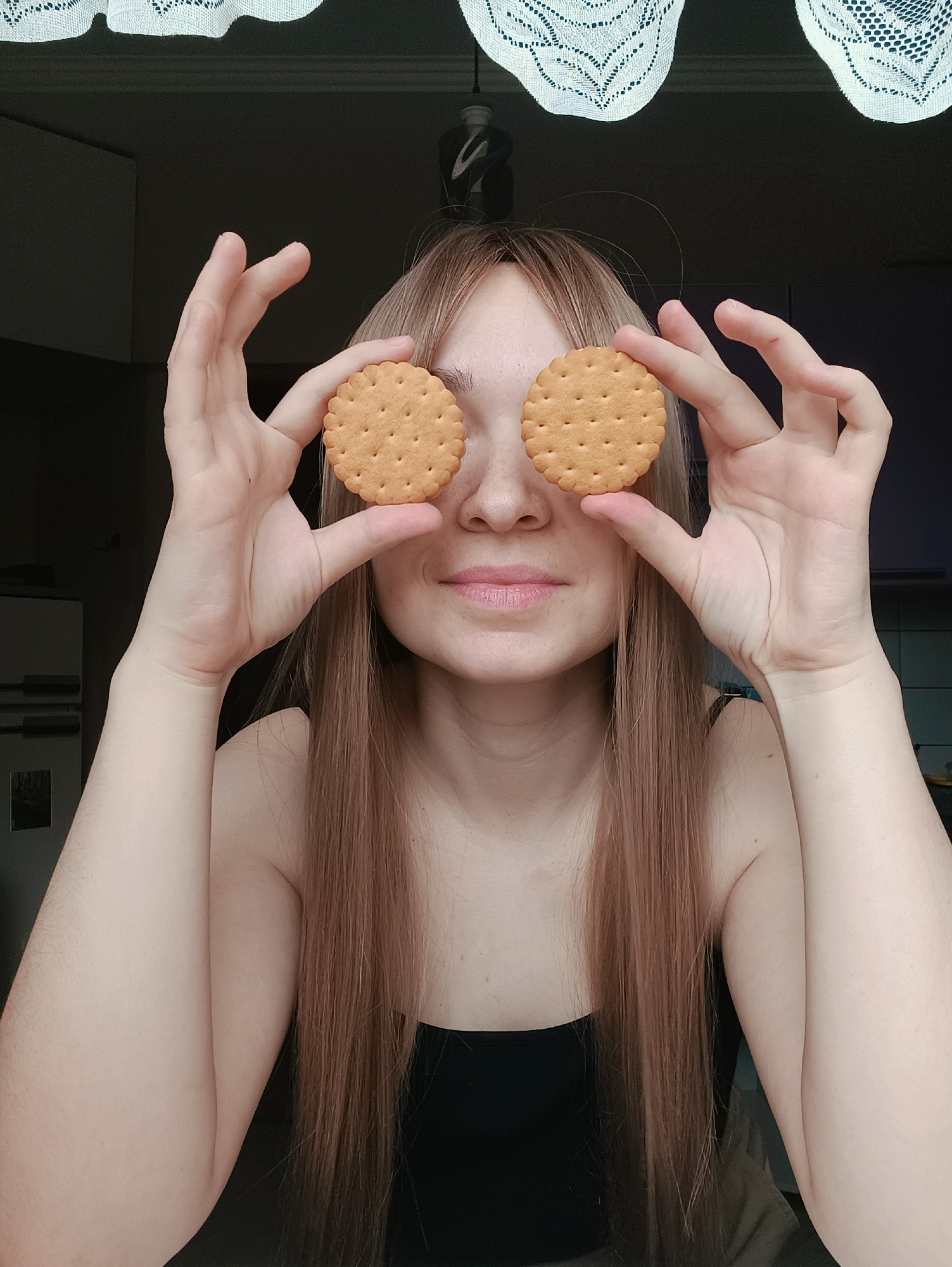 A young woman is centered in the frame, holding two round, light brown cookies over her eyes. She is looking directly at the camera and smiling slightly. Her long, light brown hair is parted in the middle and falls straight down. She is wearing a black top. The background includes a window with white lace curtains at the top of the frame, and glimpses of an interior space with light and dark areas. The photograph appears to have been taken indoors. There is no indication of it being taken in the city of Izhevsk, Russia.