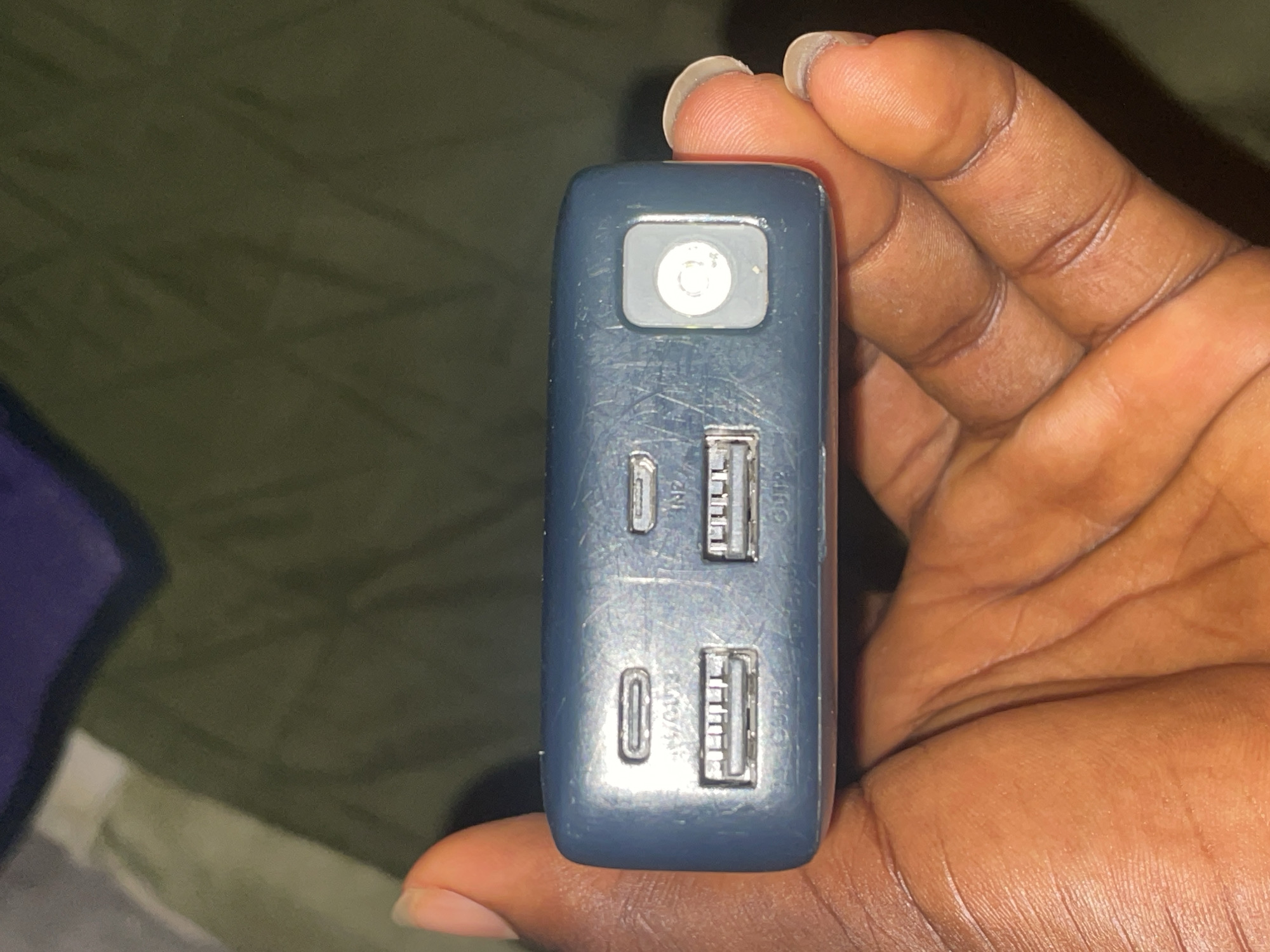 A dark blue power bank is held in a person's hand against a dark green, textured surface. The power bank has multiple ports visible: a USB-C port labeled "IN/OUT", a USB-A port labeled "OUT1", and another USB-A port labeled "OUT2". To the right of the ports is a small, circular, white button. The person's fingers and thumb are wrapped around the power bank. The background appears to be fabric with a darker purple section visible at the top. The image is taken from a top-down perspective.