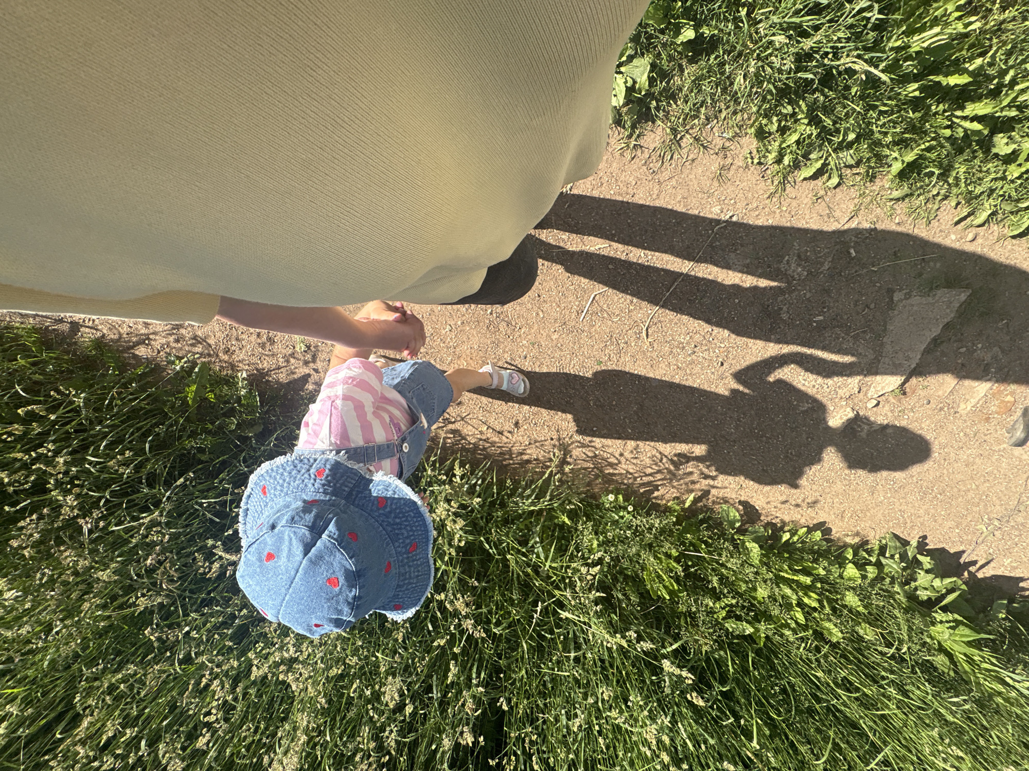 An overhead, slightly angled photograph depicts an adult and a young child walking hand-in-hand on a sunlit dirt path flanked by tall green grass. The adult, seen from the mid-torso down on the right, wears a light yellow, short-sleeved, ribbed top and dark shoes. Their right hand holds the child's left hand. The child, positioned slightly ahead and to the left, wears a denim bucket hat adorned with red heart patterns, a pink and white striped top, denim overalls, and white sandals with a pink heart detail on the left foot. The child is leaning forward, seemingly looking towards the ground or the grass. Long, distinct shadows of both figures extend onto the path in front of them, indicating bright sunlight. The path is unpaved, light brown, with dense, vibrant green grass and some lighter-colored seeding plants bordering it on both sides. The specific location within the city of Chaikovsky, Russia, is not discernible from the visual information presented in the image.