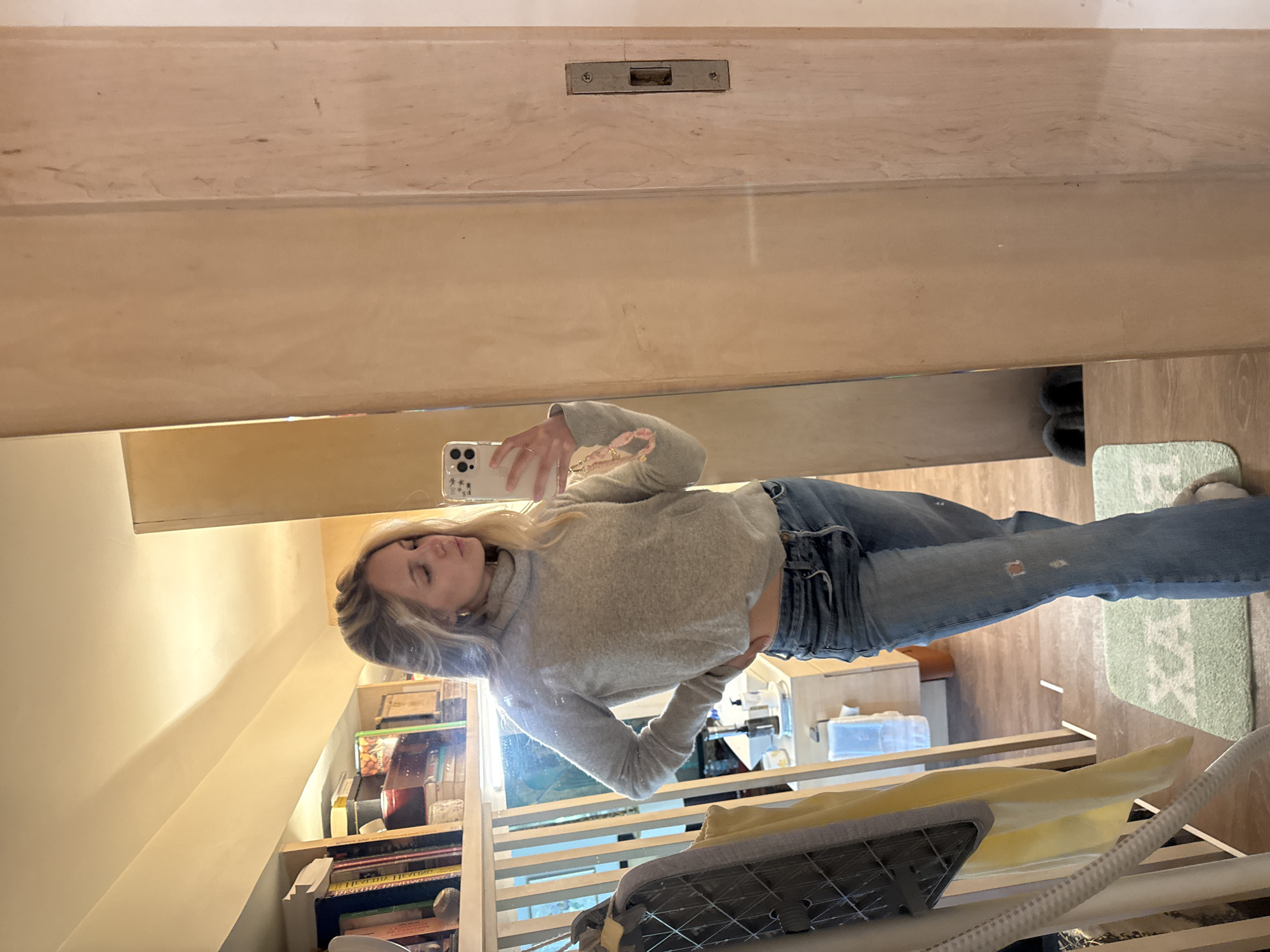 A young woman with blonde hair is taking a mirror selfie in a room. She is wearing a light gray turtleneck sweater and blue jeans. The room appears to be a home, with bookshelves on the left side of the image. There is a large mirror on the right side of the image, reflecting the woman and the room. The floor is wooden and there is a light green rug with a white pattern on it. There are also some shelves and cabinets visible in the background. The overall lighting in the room is warm and inviting. This image was taken in Mexico City, Mexico.